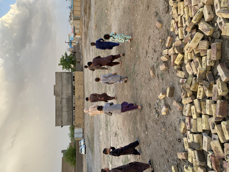 The image depicts an open, unpaved plot of land in the city of Kandahar, Afghanistan, under an overcast sky. In the foreground, a substantial pile of scattered yellowish-red bricks occupies the lower portion of the frame. Nine individuals, primarily children and young males, are observed dispersed across the dry, uneven dirt field. Most are wearing traditional shalwar kameez in varying shades of brown, grey, and blue; one smaller child, possibly female, wears a light-colored patterned outfit. Individuals are walking, with some appearing to move more rapidly than others, suggesting movement or play. In the background, an unpainted, multi-story concrete building with visible window openings stands behind a lower, sand-colored perimeter wall. Two white sedan vehicles are parked behind a metal fence structure to the left of the concrete building. Green vegetation, including a tree, is visible near the buildings. Additional urban structures and residential buildings line the distant horizon, and a flock of birds is silhouetted against the cloud-filled sky.