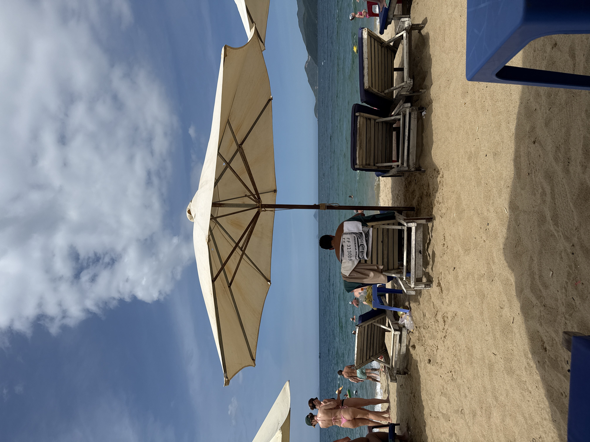 The image depicts a sunny day at a beach in Nha Trang, Vietnam, likely a popular tourist area given the presence of beach chairs, umbrellas, and numerous people enjoying the sea. In the foreground, a large, beige beach umbrella casts a shadow over the sandy beach. Several empty lounge chairs are arranged on the sand. Further back, a man sits in a chair facing away from the camera, with a towel draped over the back. To the left, two women in bikinis stand on the wet sand near the water's edge. Other beachgoers are visible in the shallow water, some swimming and others standing. The sea stretches out to the horizon, with hills or mountains visible in the distance. The sky is a vibrant blue with scattered white clouds. The overall scene conveys a relaxed and leisurely atmosphere typical of a beach resort.