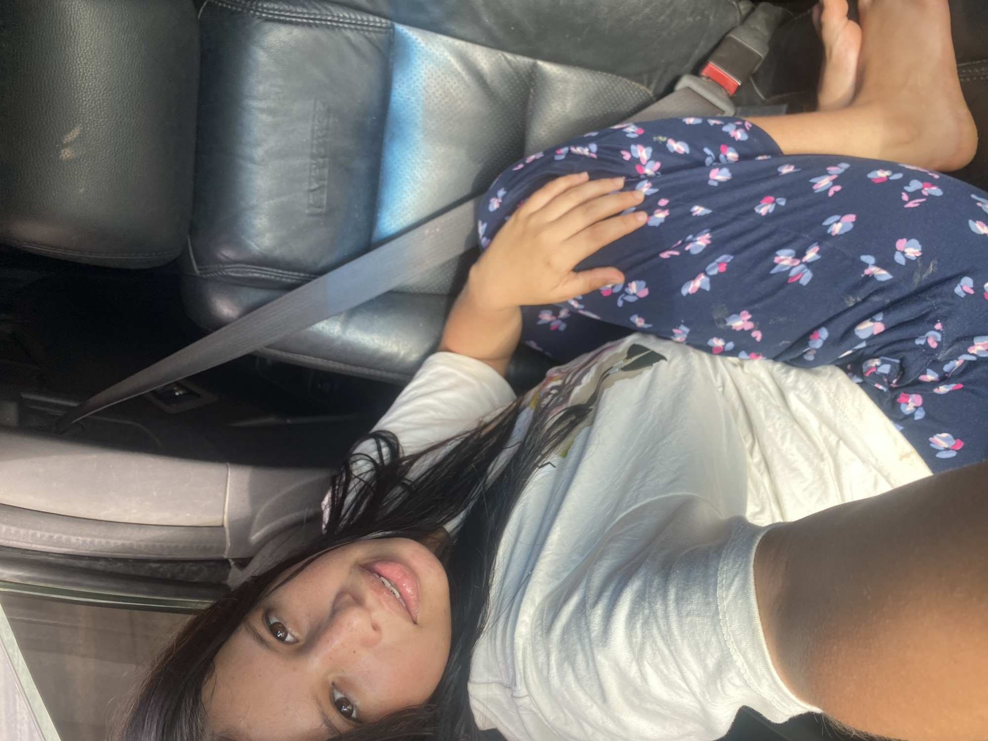 The image depicts a person lying in the passenger seat of a car. The person is wearing a white t-shirt and floral-patterned blue pants. They are positioned with their head near the car door and feet near the dashboard. The seatbelt is fastened across their midsection. The car's interior includes black leather seats. The location of the scene is not identifiable as being in Juan Griego, Venezuela, based on the visual information in the image.