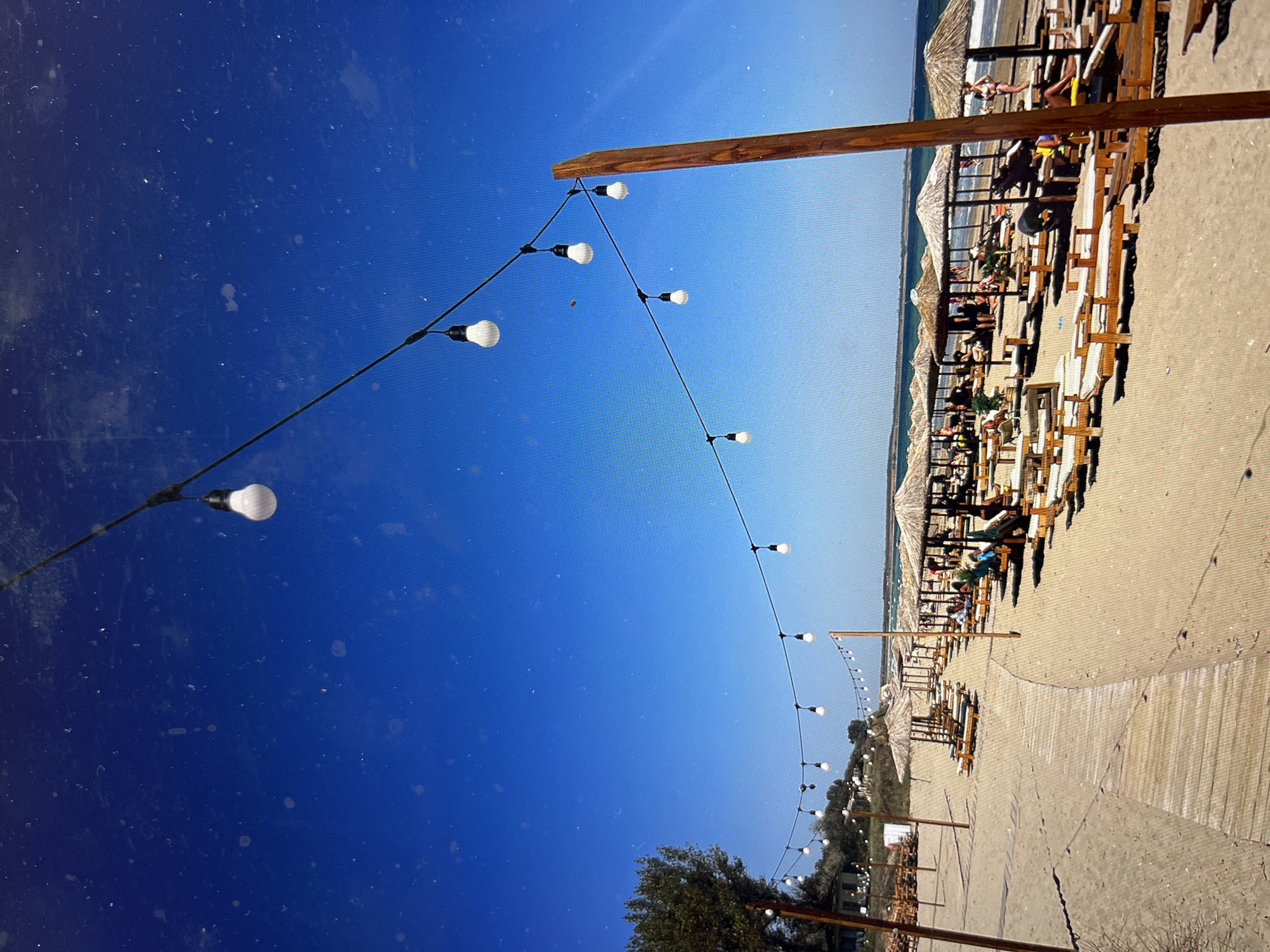 The image depicts a beach scene under a clear, blue sky. The beach is lined with sun loungers, tables, and thatched umbrellas, suggesting a tourist or recreational area. People are visible in some of the loungers, indicating they are relaxing or sunbathing. A string of lights is strung overhead, supported by wooden posts, adding to the relaxed atmosphere. The environment appears to be sunny and warm, typical of a beach location. The precise location is not specified in the image, but it appears to be a well-maintained beach resort.