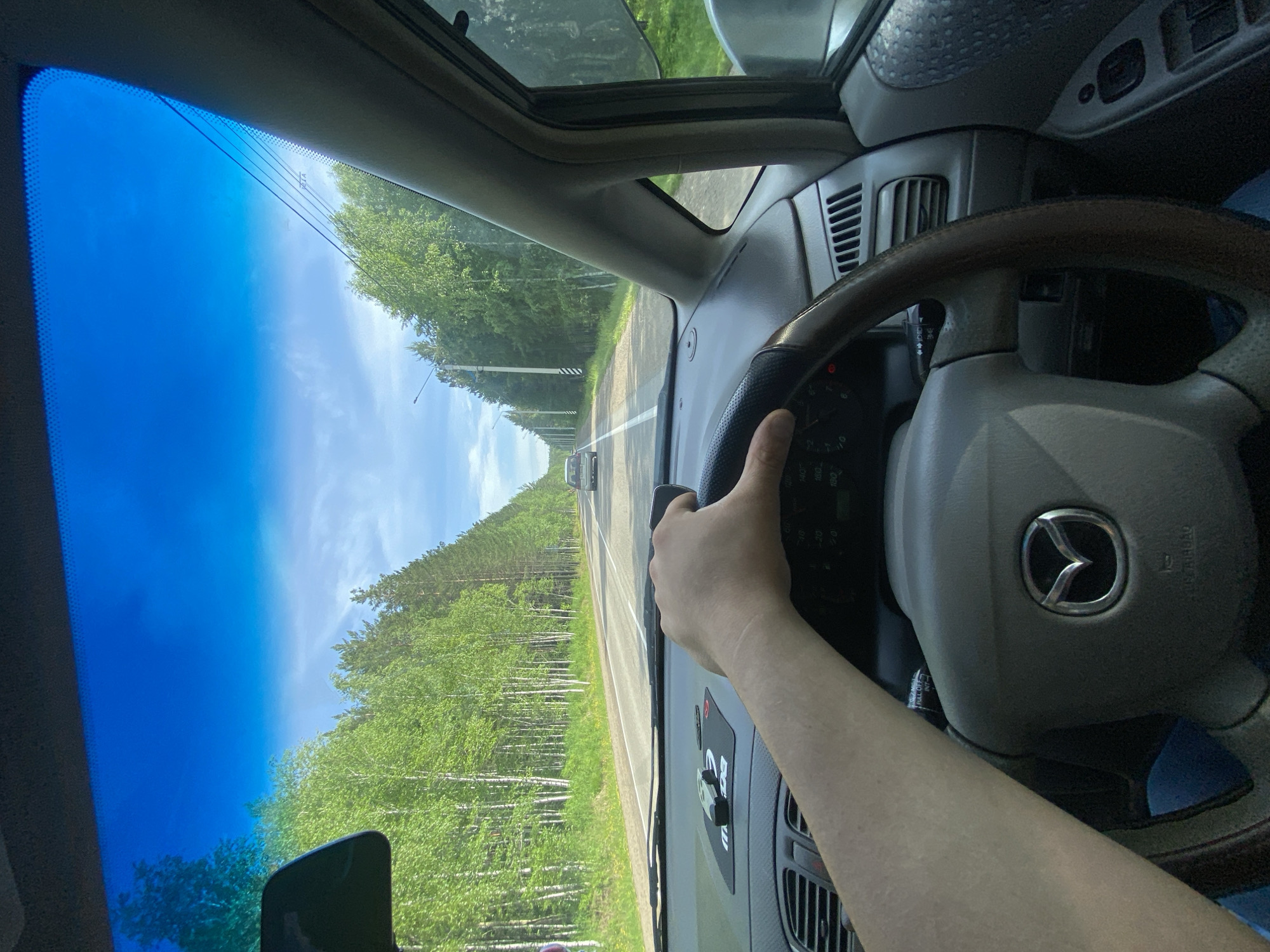 The image captures the interior of a car from the driver's perspective, with a hand on the steering wheel. The car is driving on a two-lane asphalt road through a forest. The road is lined with trees, primarily birch and pine, and there are power lines visible along the side of the road. The sky is blue with some scattered clouds. In the distance, another car can be seen driving in the same direction. The dashboard of the car is visible, including a Mazda logo on the steering wheel and a GPS device on the dashboard. The scene is consistent with a drive on a rural road in Burdakovka, Russia, during daylight hours.