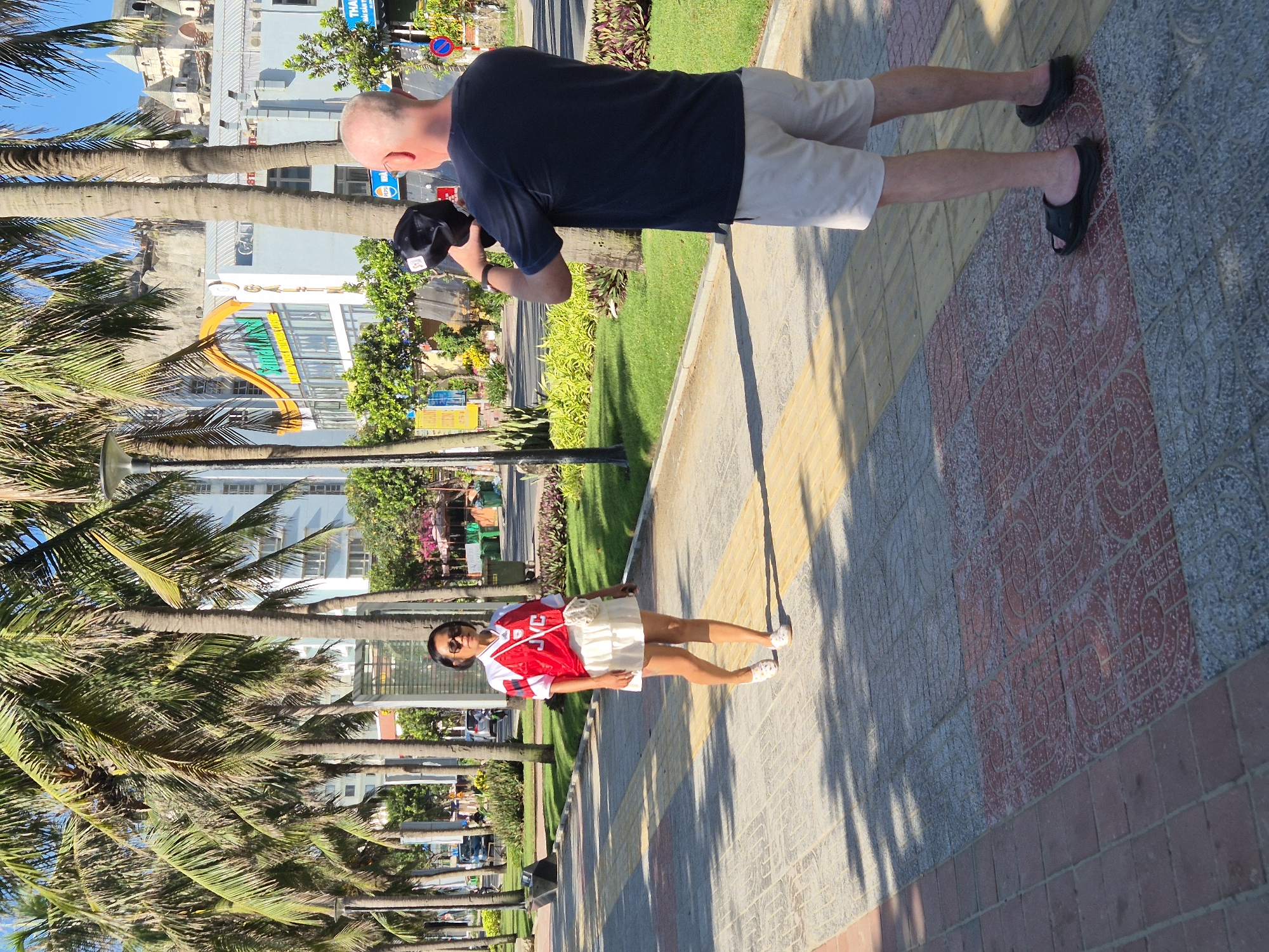 In Đà Nẵng, Vietnam, a man with a shaved head and glasses, wearing a navy blue t-shirt and beige shorts, stands on a patterned sidewalk, holding a black baseball cap. He appears to be taking a picture of a woman walking towards him. The woman, dressed in a red and white sports jersey and a white skirt, is wearing sunglasses and a small crossbody bag. She is on the same sidewalk, with palm trees lining the left side of the image. In the background, buildings with signage are visible, suggesting an urban environment. The lighting indicates it is daytime, with shadows cast on the sidewalk.