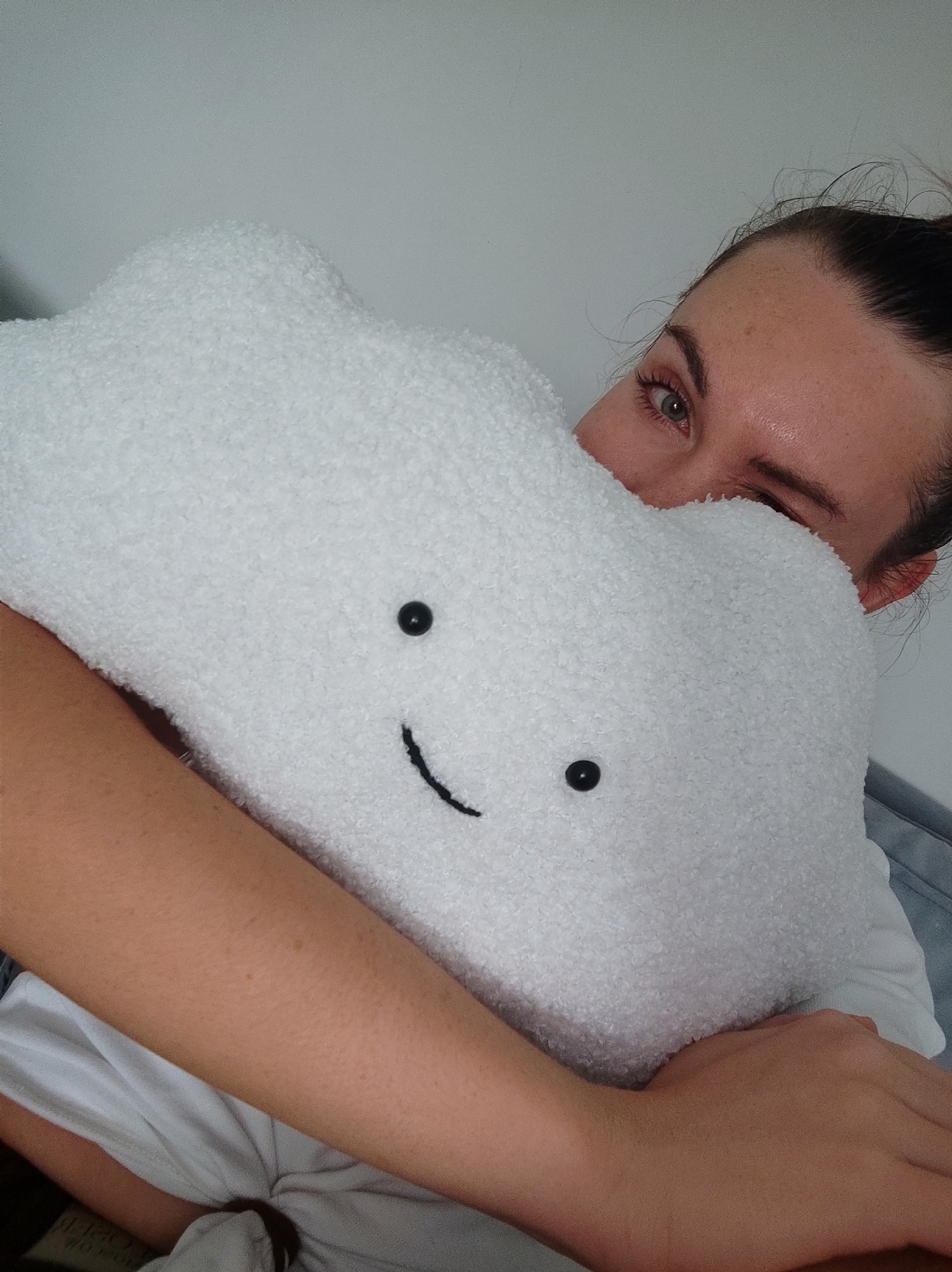 A young woman is pictured indoors, embracing a white, cloud-shaped pillow with a smiling face. The pillow features stitched black eyes and a curved mouth. The woman winks with her right eye towards the viewer. Her hair is pulled back. She is wearing a white shirt. The background consists of a white wall. There is no evidence in the image that indicates the location as Votkinsk, Russia.