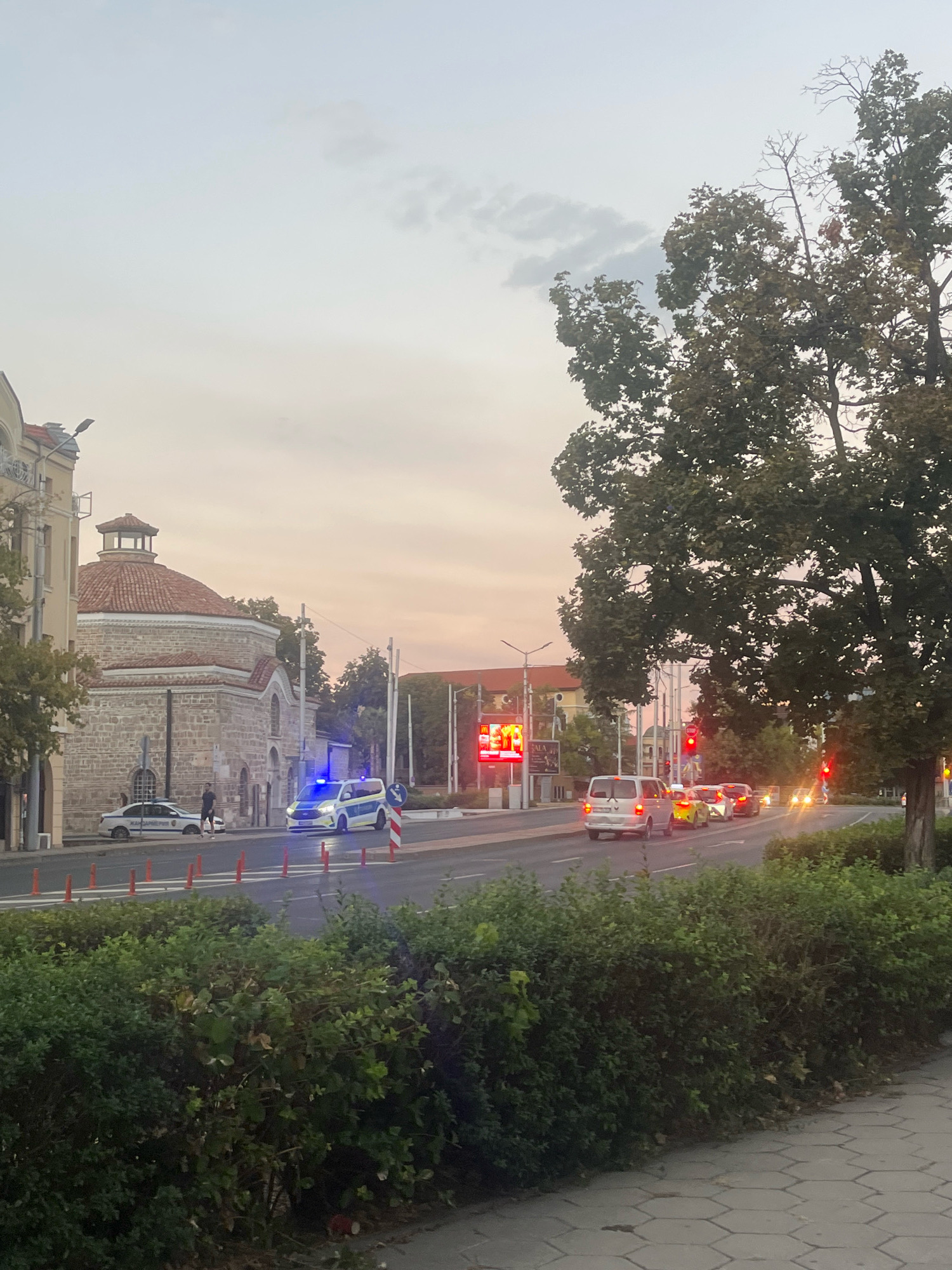 The image depicts a street scene in Plovdiv, Bulgaria, featuring a prominent historical building with a domed roof on the left. Several vehicles are visible on the road, including a police van with flashing blue lights, a police car, and other cars in traffic. A man is standing next to the police car. The sky is overcast with soft, diffused light, suggesting it is either early morning or late evening. The foreground is occupied by a dense hedge, and a paved sidewalk is visible at the bottom right. The overall atmosphere appears calm, with the presence of emergency vehicles indicating a potential incident or traffic control.