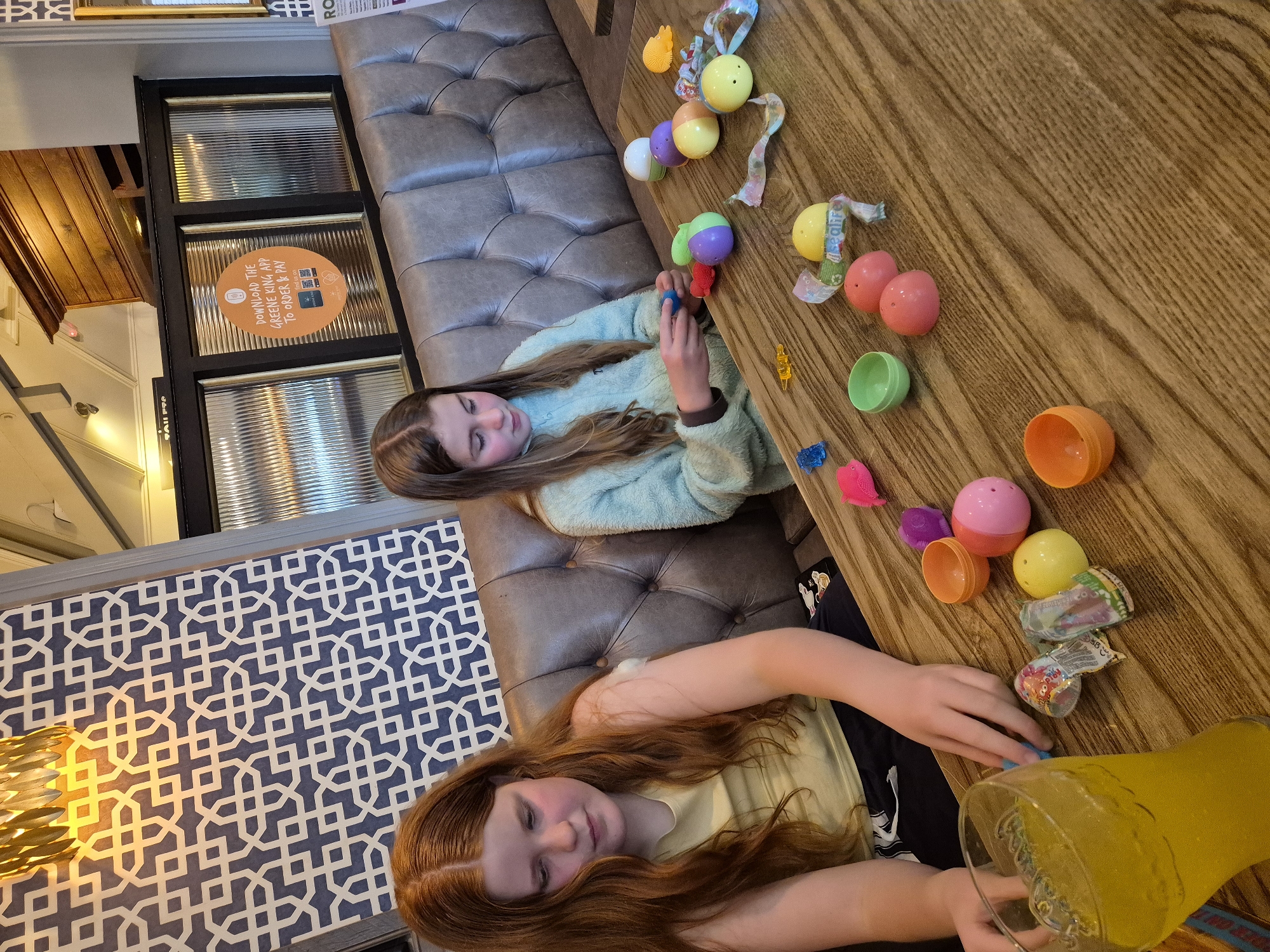 Here is a detailed description of the image:
**Scene:** An indoor setting, likely a restaurant or pub, with two young girls at a wooden table.
**People:**
* Two girls: One has light brown hair and is wearing a light blue sweater. The other has red hair and is wearing a light yellow shirt. They appear to be related, possibly sisters.
**Objects:**
* Plastic Easter eggs of various colors (pink, yellow, orange, green, purple, white).
* Small toys or candies, some still wrapped.
* A large pitcher of a yellow beverage, possibly lemonade or juice.
* A brown leather upholstered banquette or booth seating.
* Decorative wall with a repeating geometric pattern in blue and white.
* A glass window panel with frosted glass.
* A sign with orange background is attached to the glass window panel saying "DOWNLOAD THE GREENE KING APP TO ORDER & PAY".
**Actions:**
* The girls are playing with the plastic Easter eggs and their contents, possibly sorting or organizing them.
* The girl with the red hair is reaching towards or pouring from the pitcher of yellow beverage.
**Interactions:**
* The girls appear engaged in a shared activity, possibly involving Easter-themed toys and treats.
**Environment:**
* The background shows elements of a commercial establishment, such as the patterned wall, the window, and the sign.
**Location Context:** The sign mentioning "GREENE KING APP" suggests this is a Greene King pub or restaurant, which is a popular chain in the United Kingdom. This image was taken in Breckland District, United Kingdom.