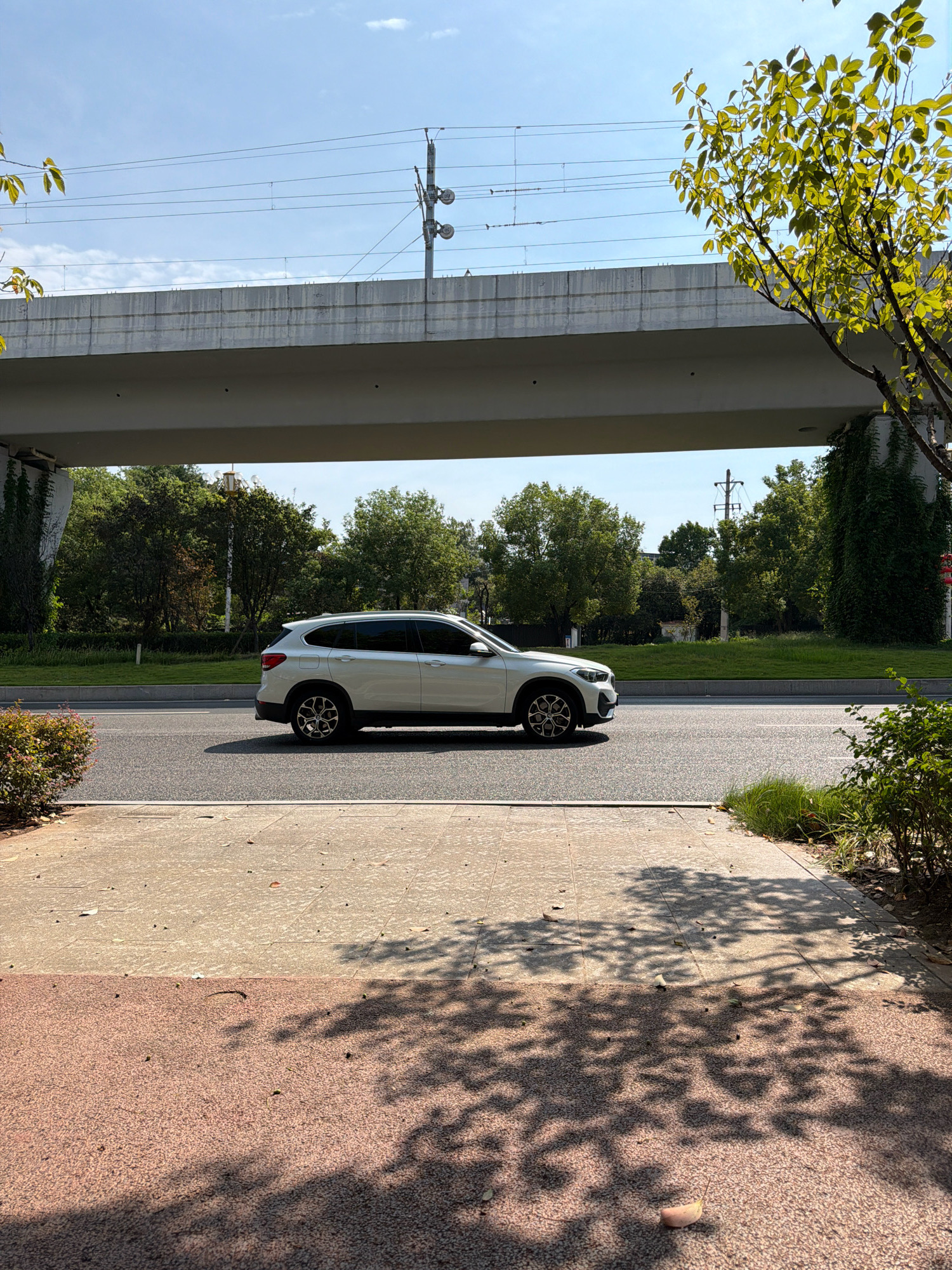 A white SUV is driving on an asphalt road under an elevated railway track structure in Jiangjin, China. The sky is clear and blue with some clouds. Trees and greenery are visible on either side of the road, with a sidewalk in the foreground. The scene is captured from a slightly elevated perspective, looking across the road towards the trees.