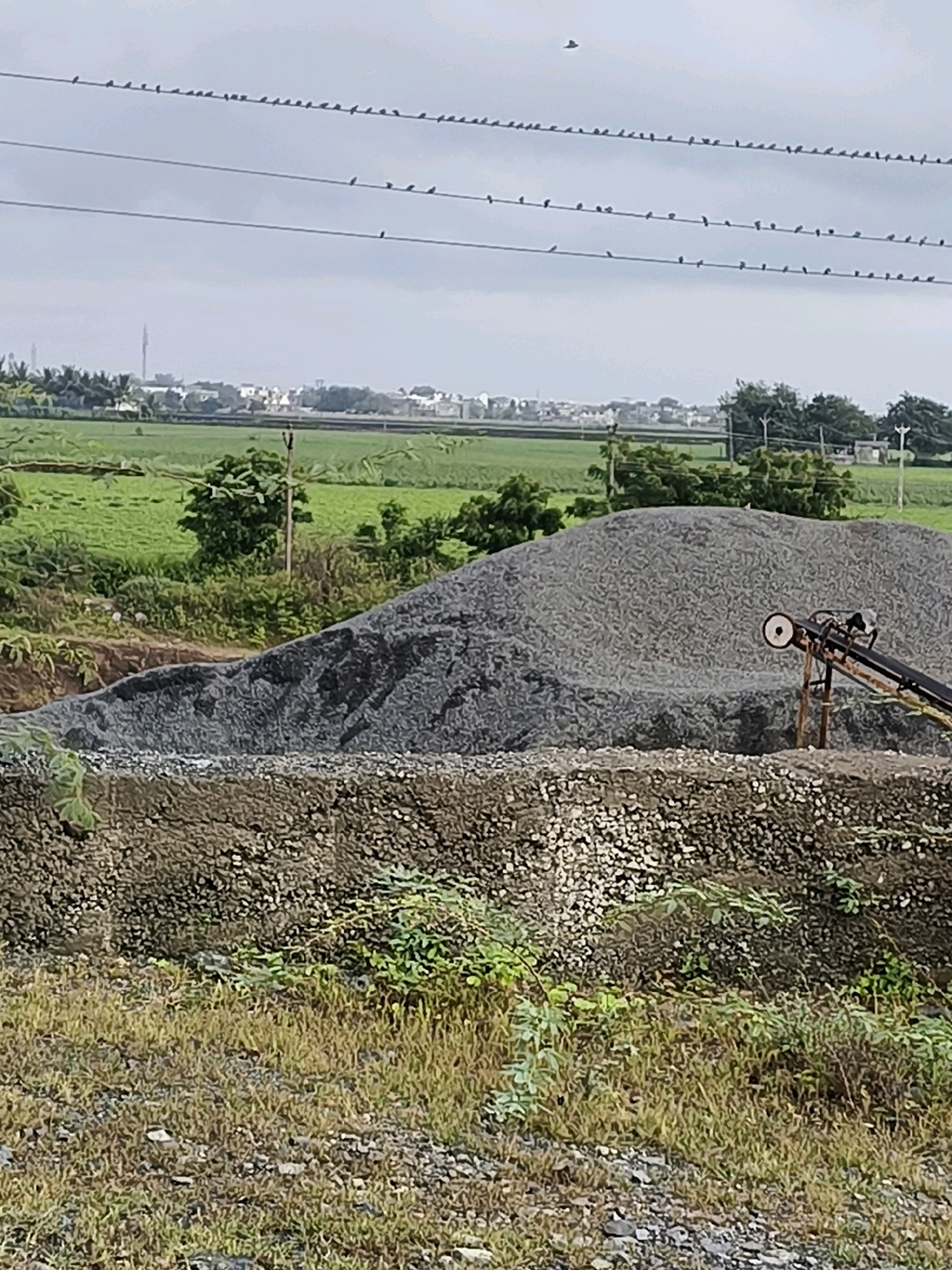 This is a medium-distance, eye-level, outdoor photograph taken in Mahobat Para, India. The scene is dominated by a large pile of grey gravel in the foreground and midground. A conveyor belt system, with a visible wheel and motor, is positioned on the right side of the gravel pile. Behind the gravel, there is a green field with some trees. In the distance, a town or city with buildings is visible, partially obscured by trees. The sky is overcast with a few birds silhouetted against it. Power lines with numerous birds perched on them are stretched across the upper part of the frame. The overall impression is of an industrial or construction site on the outskirts of a settlement.