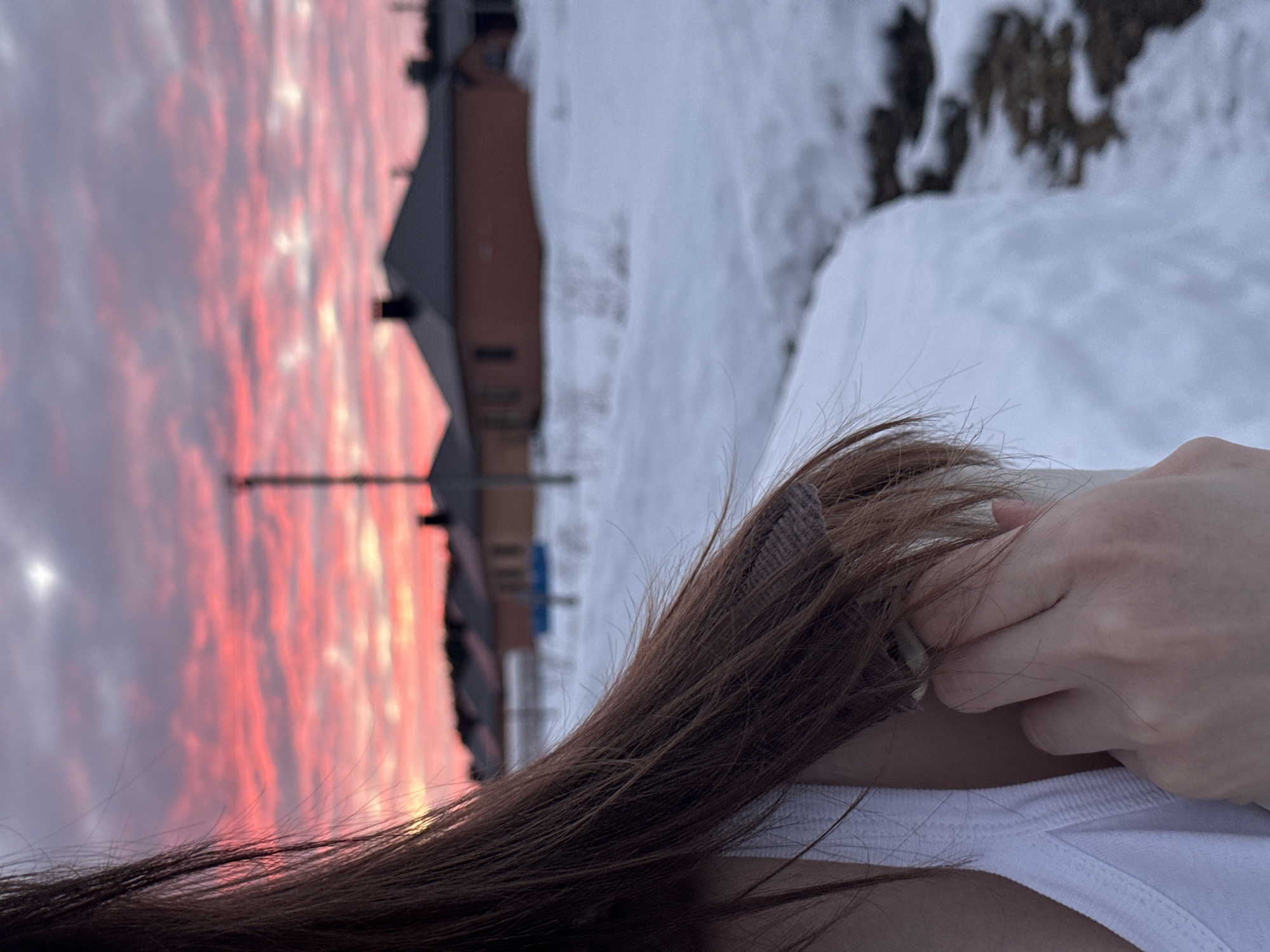 The image displays a close-up, over-the-shoulder perspective of an individual positioned on the left side of the frame. The subject has long, dark brown hair and is wearing a white tank top. Their right hand is visible near their neck, partially obscured by hair, with a light-colored hair clip present in the hair. The background, identified as a residential area in Chaikovsky, Russia, features a snowy landscape under a dramatic twilight sky. The ground is covered in white snow, intermingled with patches of dark, exposed earth, indicating melting. In the midground, several single-story houses with brown walls and dark roofs are visible in a row, along with a tall, slender utility pole. The sky above is illuminated with vivid pink and red clouds, characteristic of either a sunrise or sunset, transitioning to lighter purplish-grey tones at the top.