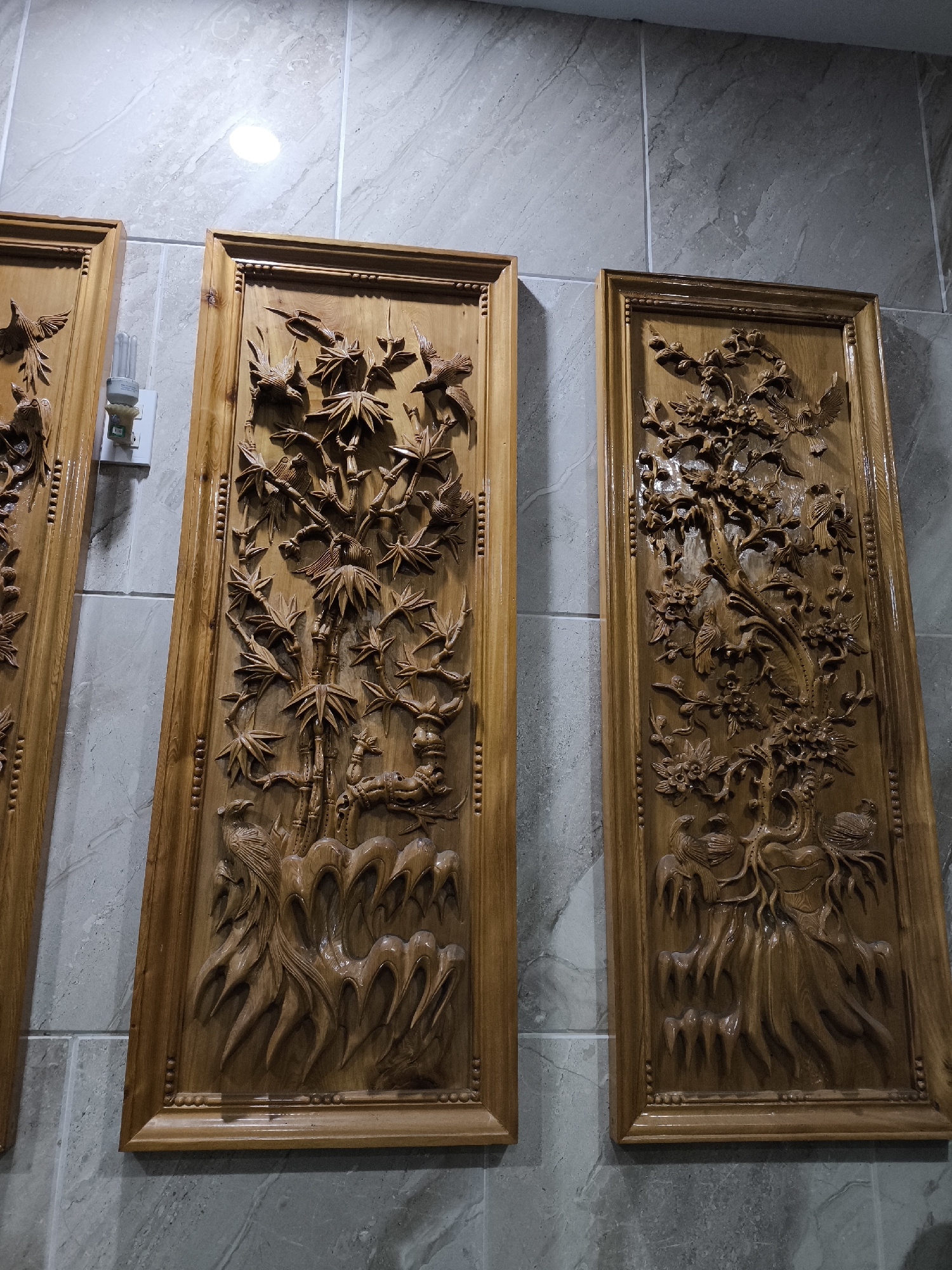 This image depicts three framed wood carvings displayed on a gray tiled wall. The carvings, made of a light-colored wood with a glossy finish, feature intricate designs of birds and natural elements such as bamboo and trees with flowers. The carving on the left is partially visible, and the two fully visible carvings are oriented vertically. The carving in the center depicts bamboo stalks and numerous birds in flight and perched. The carving on the right showcases a tree with flowers and birds, and at the bottom, what appears to be a phoenix-like bird. A light fixture is visible on the wall between the leftmost and center carvings. The wall is made of rectangular gray tiles with visible grout lines. The lighting in the scene is overhead and appears to be from a ceiling light, casting a bright illumination in the upper portion of the image. The overall setting suggests an interior space, possibly a home or a gallery, in Nha Trang, Vietnam.