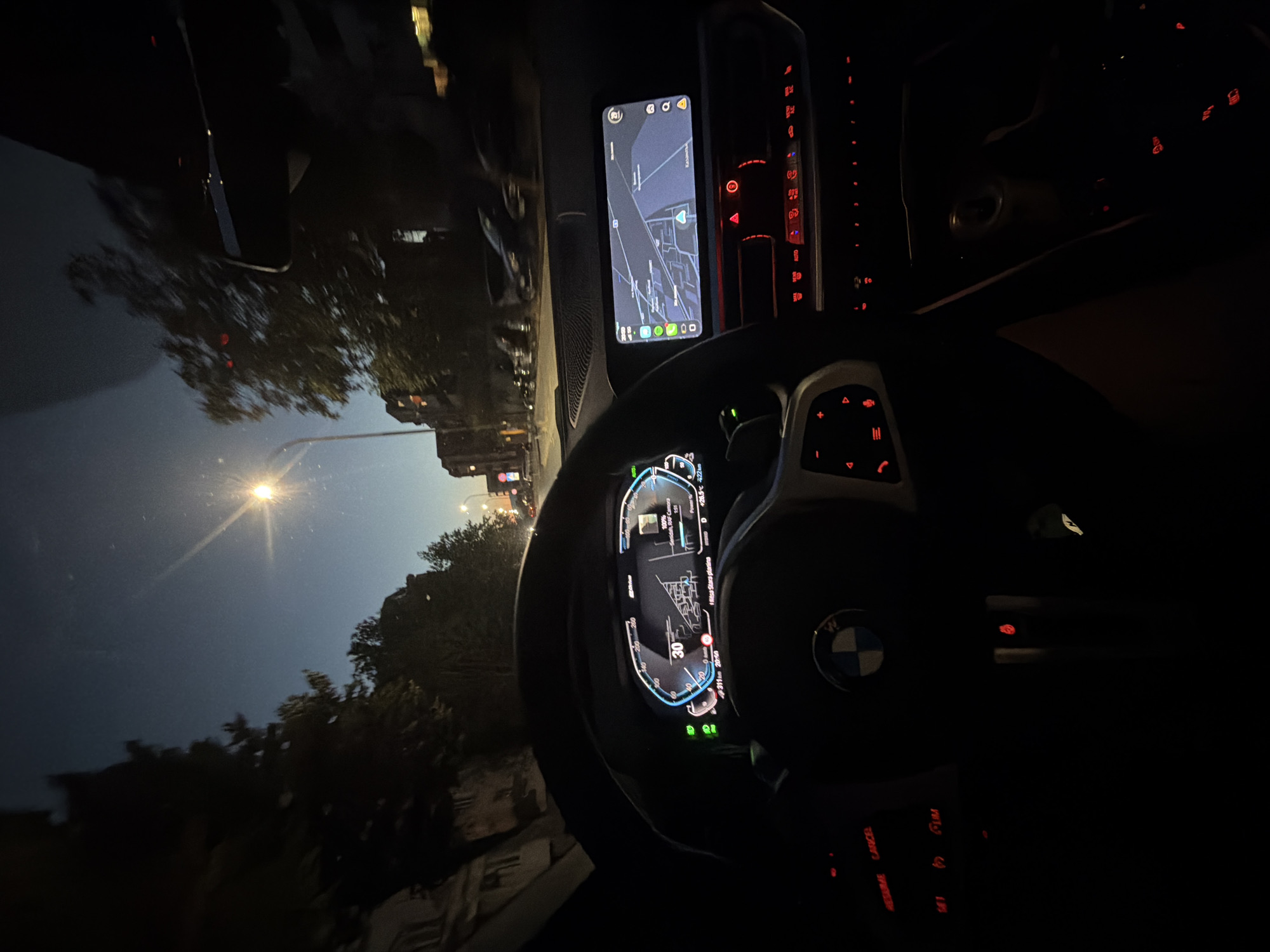 This is a first-person perspective shot from inside a car at dusk, possibly in the city of Kazanlak, Bulgaria. The driver's dashboard is illuminated, showing a digital speedometer indicating 30 km/h, along with navigation displayed on a central infotainment screen. The steering wheel, with the BMW logo, is visible. Red illuminated buttons and controls are present. Outside, streetlights and buildings are visible in the twilight, with trees lining the road. The car appears to be in motion on a city street.