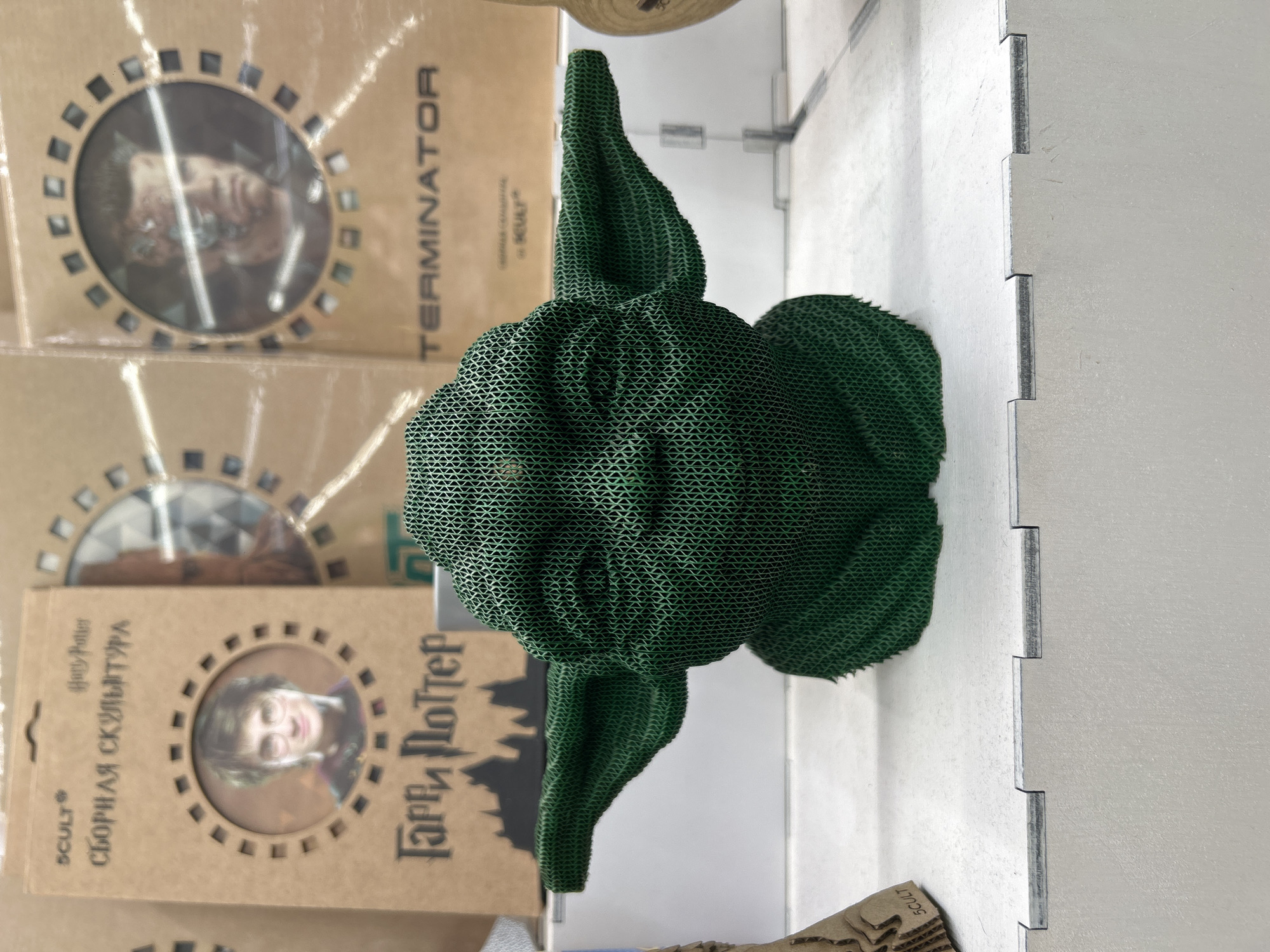 The image shows a green Yoda sculpture, possibly 3D printed, standing on a shelf. To the left of Yoda, there are two cardboard boxes: one labeled "Terminator" and the other "Harry Potter" in Russian (Гарри Поттер). "SCULT" and "СБОРНАЯ СКУЛЬПТУРА" (Assembly Sculpture) are also visible on the Harry Potter box. The background and shelving appear to be made of white interlocking panels. The location is Chaikovsky, Russia, as indicated in the prompt.