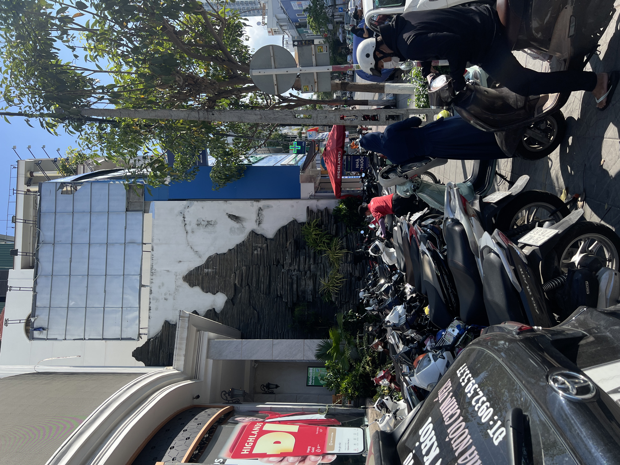 The image captures a bustling street scene in Nha Trang Ward, Vietnam. Numerous parked motorbikes line the sidewalk, creating a dense and chaotic parking area. Several individuals are visible around the motorbikes, some wearing helmets, which indicates their use as transport. The vehicles obscure much of the sidewalk and street behind them.
A building is situated behind the motorbikes, with a distinctive decorative wall element resembling dark stone or rock formations. The building appears to house commercial establishments; a "Highlands Coffee" advertisement is visible on the storefront.
Street signage, trees, and other buildings are apparent in the background, confirming the urban setting. The overall environment is typical of a busy commercial district, with a high density of traffic and commercial activity.