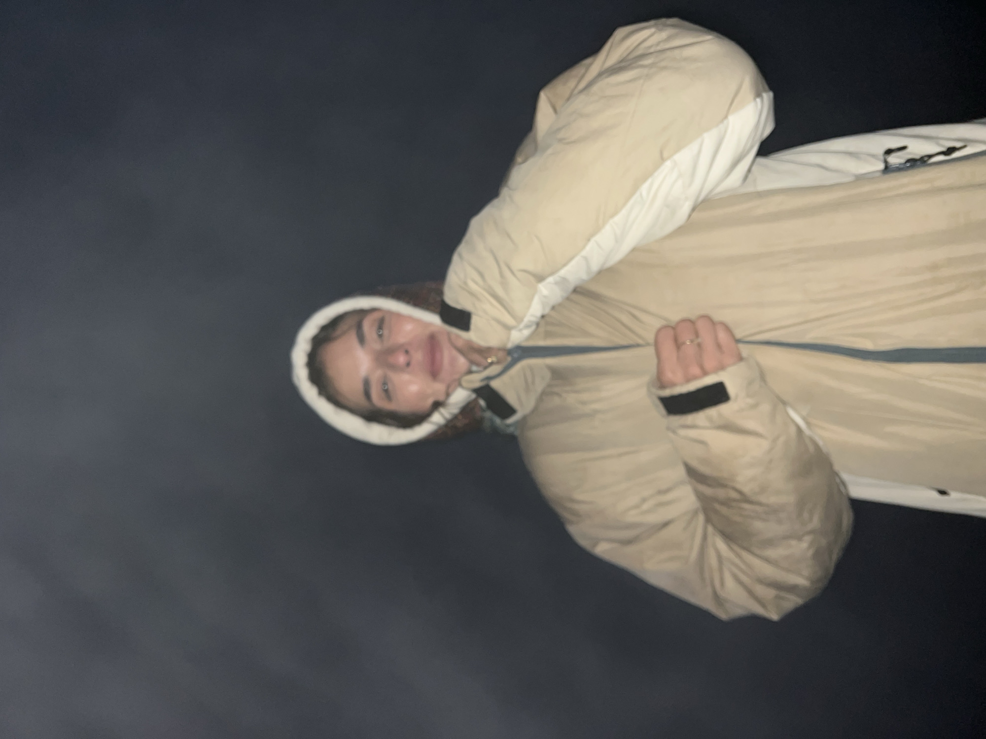 A young woman with medium-length brown hair is wearing a light tan puffer jacket with a white lining around the hood and collar. She has the hood pulled up and is holding it with her hands, which are clasped in front of her chest. She is wearing a ring on her right hand. Her facial expression is one of mild discomfort or cold. The background is dark and indistinct, suggesting an outdoor environment at night, possibly in Kintamani, Indonesia, given the mention of the location. The lighting is dim, with some of the woman's face and jacket illuminated, creating a hazy effect.
