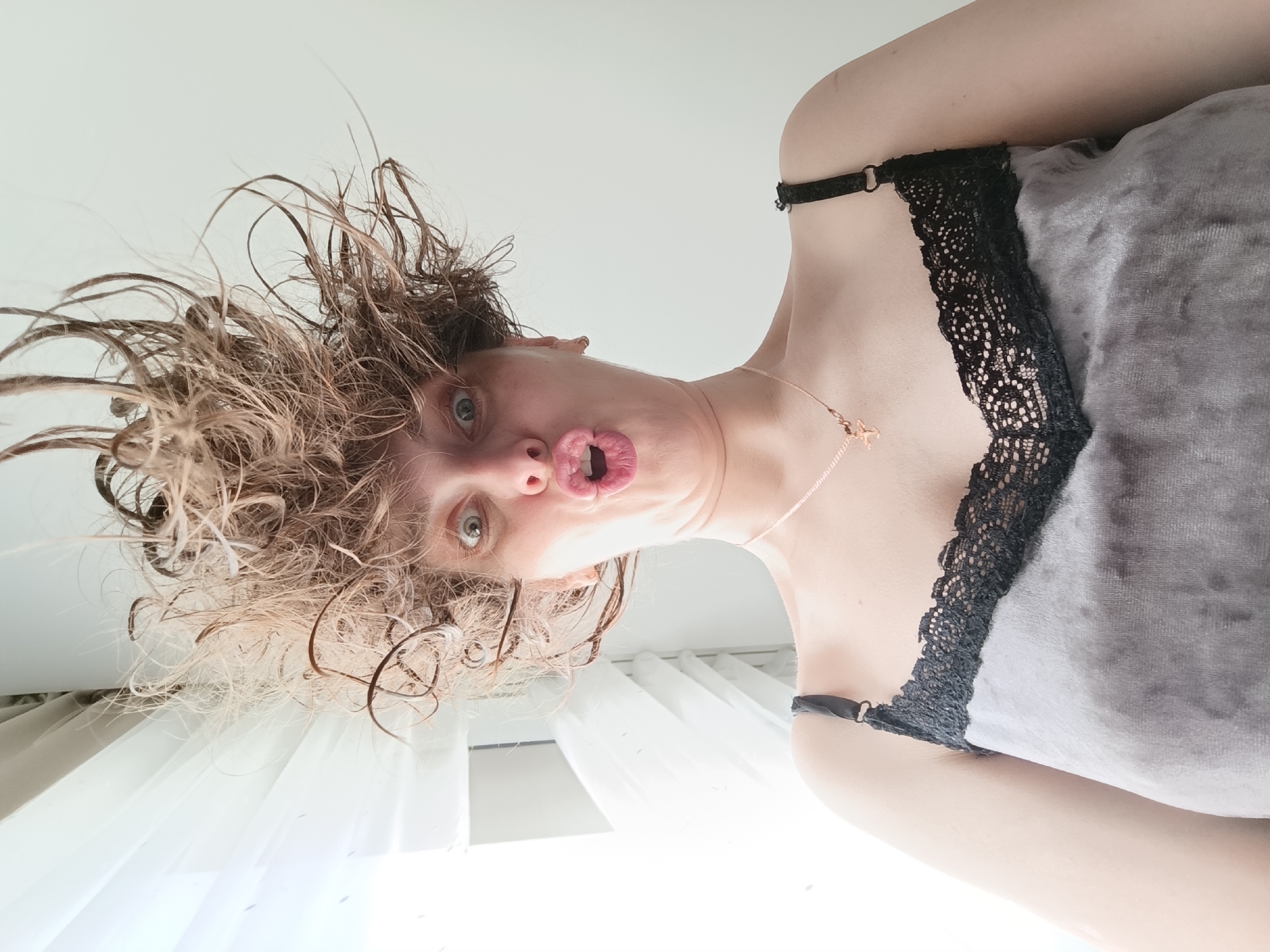 The image shows a person with damp, messy brown hair, making a pouting expression. The person is wearing a gray top with black lace trim. A thin, delicate necklace is visible. The background appears to be a white or lightly colored interior, with a glimpse of what could be a window with sheer curtains. The perspective is from below, looking up at the person. There is no explicit information that points to the city of Chaikovsky, Russia.