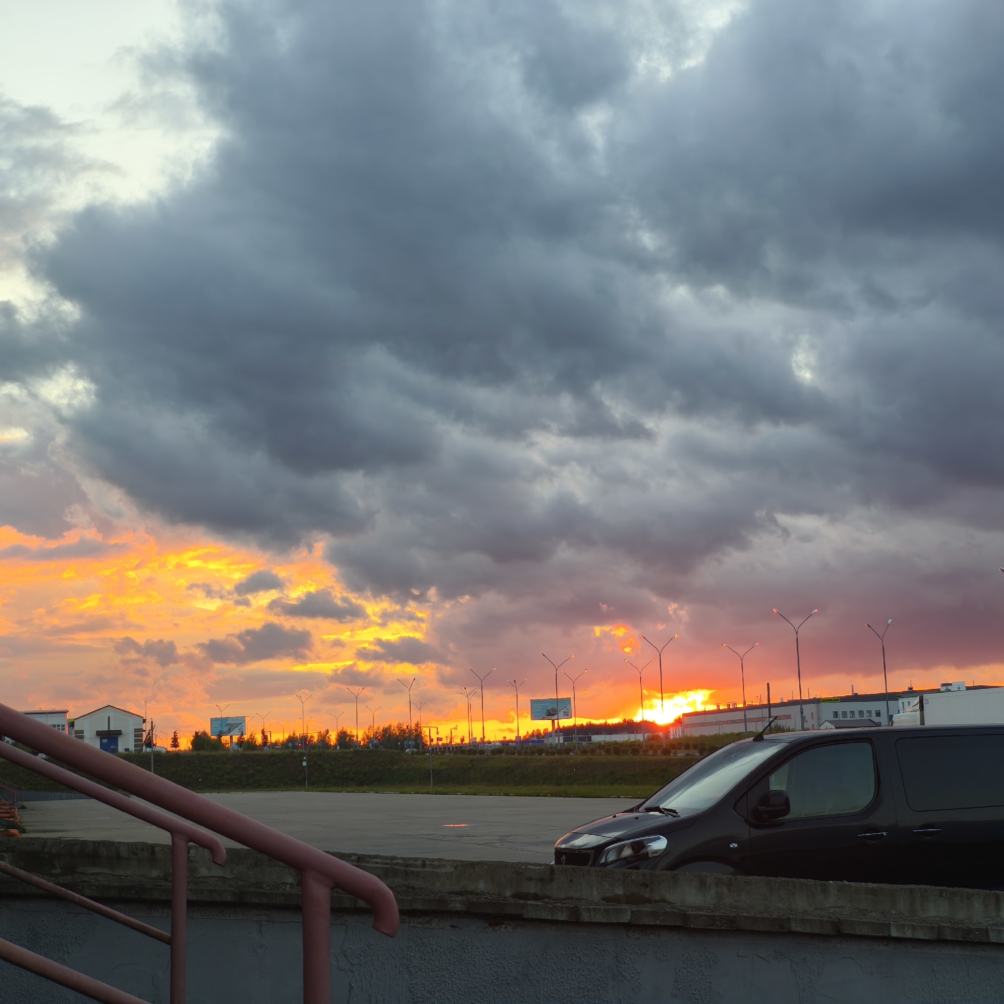 A dark-colored van is parked in an outdoor paved area under a dramatic sky filled with dark, heavy clouds, illuminated by a vibrant sunset. The sunset casts intense orange and yellow hues across the lower portion of the sky, contrasting sharply with the dark, ominous clouds above. In the background, there are several buildings, including what appears to be a warehouse or industrial facility, a smaller white building with a blue door, and other structures. Rows of streetlights with distinct lamp heads are visible along a slightly elevated area behind the paved lot, lining a strip of green vegetation and trees. A concrete barrier and a red railing are in the foreground, suggesting a walkway or elevated viewpoint. The scene appears to be an industrial or commercial zone on the outskirts of Драчкаўскі сельскі Савет, Belarus. No people are visible in the image.
