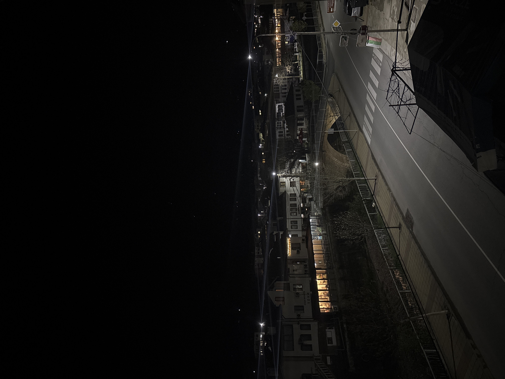 This nighttime image, taken from a high vantage point, depicts a street and surrounding buildings in Zlatograd, Bulgaria. The original image is rotated 90 degrees clockwise, showing the sky on the left and the street on the right.
The scene captures a quiet town after dark. A paved road, marked with a prominent white crosswalk and a double yellow line, stretches diagonally across the right side of the frame. Streetlights illuminate sections of the road and sidewalk, creating pools of light and casting faint streaks across the dark sky. A dark-colored car is parked on the far side of the road near a cluster of traffic signs and a light pole. The sidewalk running alongside the road features patterned paving stones in shades of yellow and grey.
On the opposite side of the road, various multi-story buildings line the street. Many windows are illuminated with warm light, suggesting occupied residences or businesses. One building clearly displays "ХОТЕЛ ТОНИ" (Hotel Toni) and "МАГАЗИН" (Shop) in Cyrillic script. In the middle ground, a arched stone structure, possibly part of a bridge or an underpass, is visible. Beyond the immediate foreground, a dense collection of buildings with scattered lights extends into the distance.
The sky, which occupies a significant portion of the left side of the image, is dark, with a few faint stars visible, indicating clear weather. Sparse, bare trees and bushes line the street and appear between buildings, characteristic of a colder season or dormant vegetation. No people or active events are visible, contributing to a tranquil and still atmosphere. Visible text includes Cyrillic on road signs, such as "ХОТЕЛ ТРАКИЯ" (Hotel Trakiya), "РИТЕЦ" (Ritetz), and "ТРЪН" (Tran) on a directional sign, as well as the building signage mentioned. A white sign with a green banner is also present but its text is indistinct.