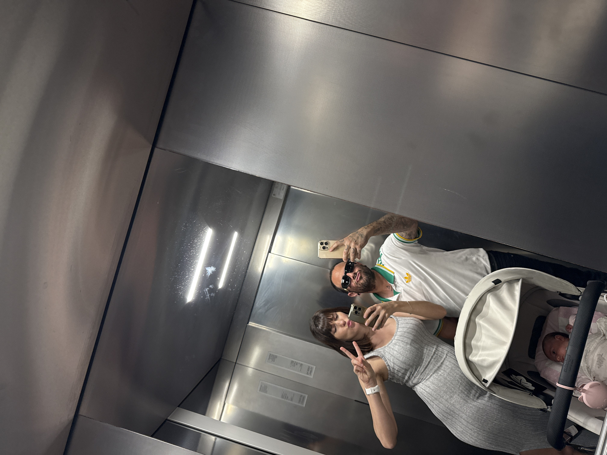 A man, a woman, and a baby are in an elevator, possibly in Pomezia, Italy. The woman holds up two fingers in a peace sign, while the man holds a phone taking a picture of their reflection in the elevator's metal walls. The baby is in a stroller. Both the man and the woman are using cell phones, and the woman is wearing a watch.