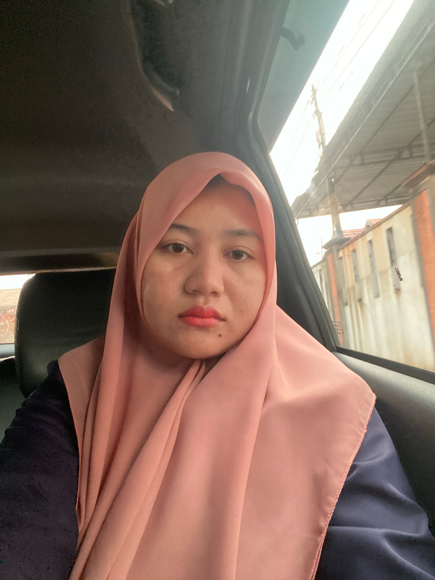 A young Indonesian woman wearing a pink hijab and a navy blue shirt is sitting in the backseat of a car. Her face is angled towards the camera, and she has light makeup on, including pink lipstick. The car's interior, including a dark leather seat, is visible. Outside the car window, a blurred background shows buildings and utility poles, suggesting an urban or suburban setting. The lighting indicates it is daytime. The image is a selfie taken from a low angle.