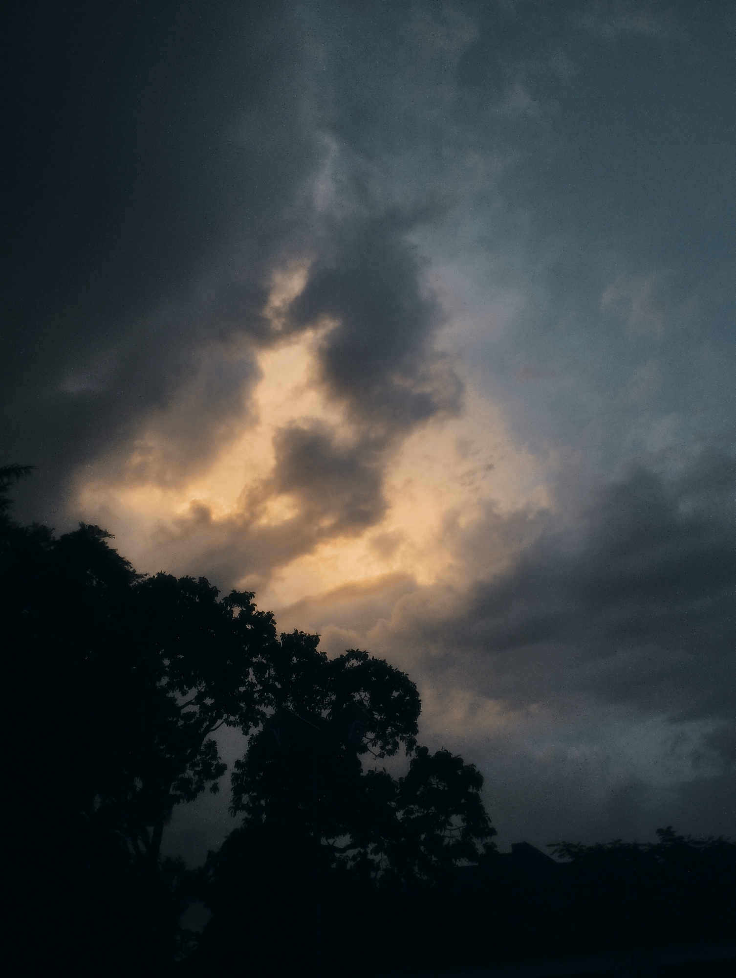 The image depicts a dark, stormy sky over a silhouetted treeline. The clouds are heavy and ominous, with a patch of warm, golden light breaking through in the upper center of the frame. The bottom portion of the image is dominated by the dark shapes of trees and possibly some low-lying structures, suggesting a natural or semi-rural setting. There are no people visible in the image. The overall mood is one of impending weather change. This scene could be observed anywhere with such atmospheric conditions, including Jos, Nigeria.