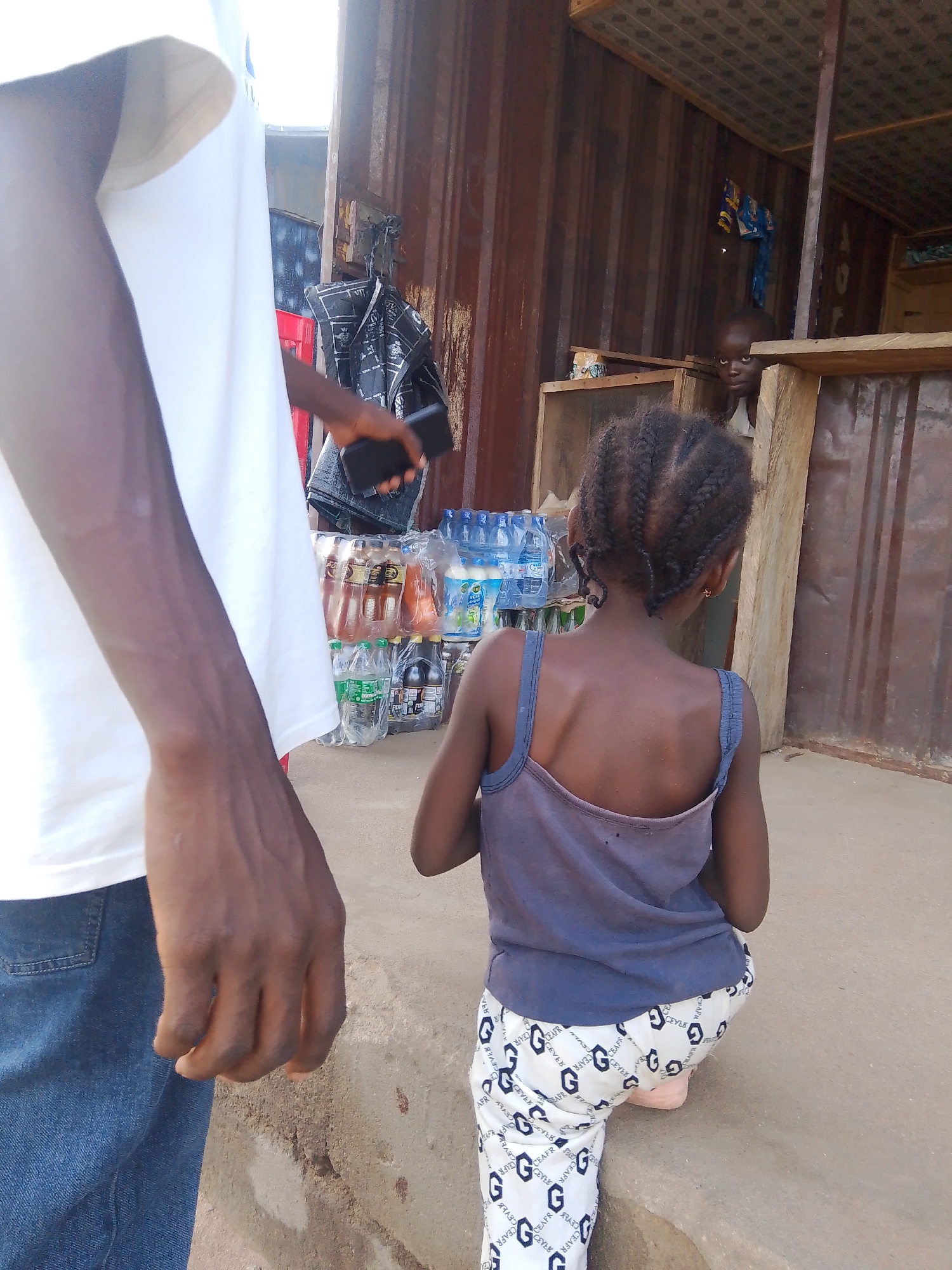 In Jalingo, Nigeria, a scene unfolds at an outdoor market stall constructed from corrugated metal. The foreground is dominated by the arm and torso of an adult, clad in a white t-shirt and blue jeans, holding a dark-colored mobile phone. To the right, a young girl, with her back to the camera, wears a dark grey tank top and white patterned pants. Her hair is styled in braids.
Behind the child, stacked bottles of beverages, including water and what appear to be soft drinks, are neatly arranged on shelves. A young boy can be seen peeking from behind a wooden counter in the background, his gaze directed towards the camera. The overall setting suggests a bustling, everyday commercial environment. The time of day appears to be daytime, with diffused sunlight indicating possibly overcast conditions or shade from a structure. The image captures a candid moment, possibly a transaction or a pause in activity at the market. The visible text on the girl's pants is a repeating pattern of "G" and other characters like "CEAFR", "CEVEK", and "AFR".