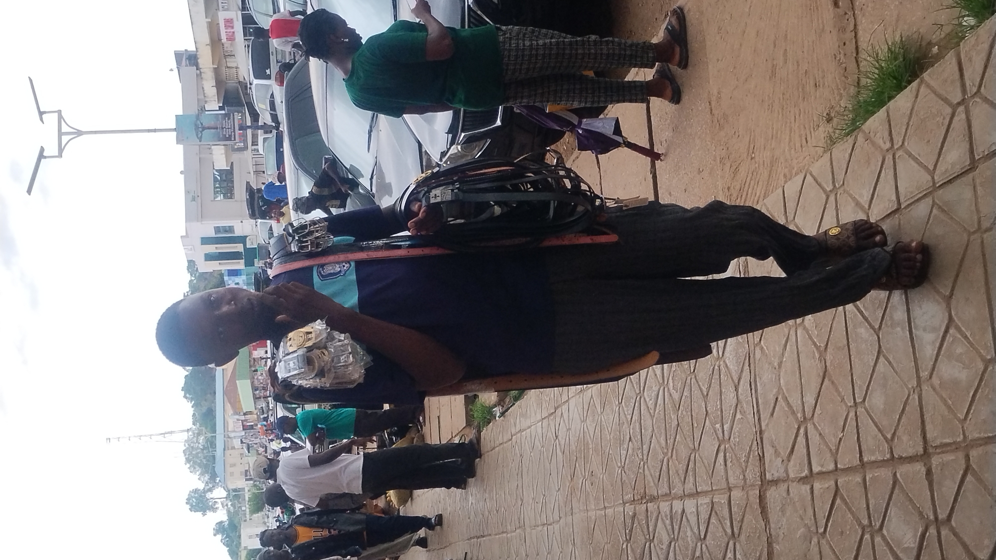 The image captures a daytime street scene, likely in an urban environment, under an overcast sky. In the foreground, a man, possibly a street vendor, stands on a sidewalk paved with geometric-patterned tiles, looking directly at the camera. He wears a dark blue and light blue polo shirt, dark pants, and brown sandals. He appears to be displaying various items for sale around his neck and chest, including small electronic devices or accessories in clear plastic packaging, a coiled black cable or hose, and metallic objects resembling car parts.
To the right and further back, another person is visible, primarily from behind, wearing a green t-shirt, patterned trousers, and dark sandals. This individual stands near a dark-colored parked car. A small purple umbrella is resting on the ground nearby. Several other vehicles are parked along the street, and various pedestrians can be seen in the background, including one wearing a white t-shirt and another in a yellow and black jacket.
The background features multiple buildings, some with white and blue facades. A tall street light pole with an attached billboard is prominent in the middle ground; the word "COMPANY" is partially visible on the billboard. On a distant building, the text "C.T.D" is discernible on a sign. The scene conveys a sense of everyday activity in a busy street market or commercial area.