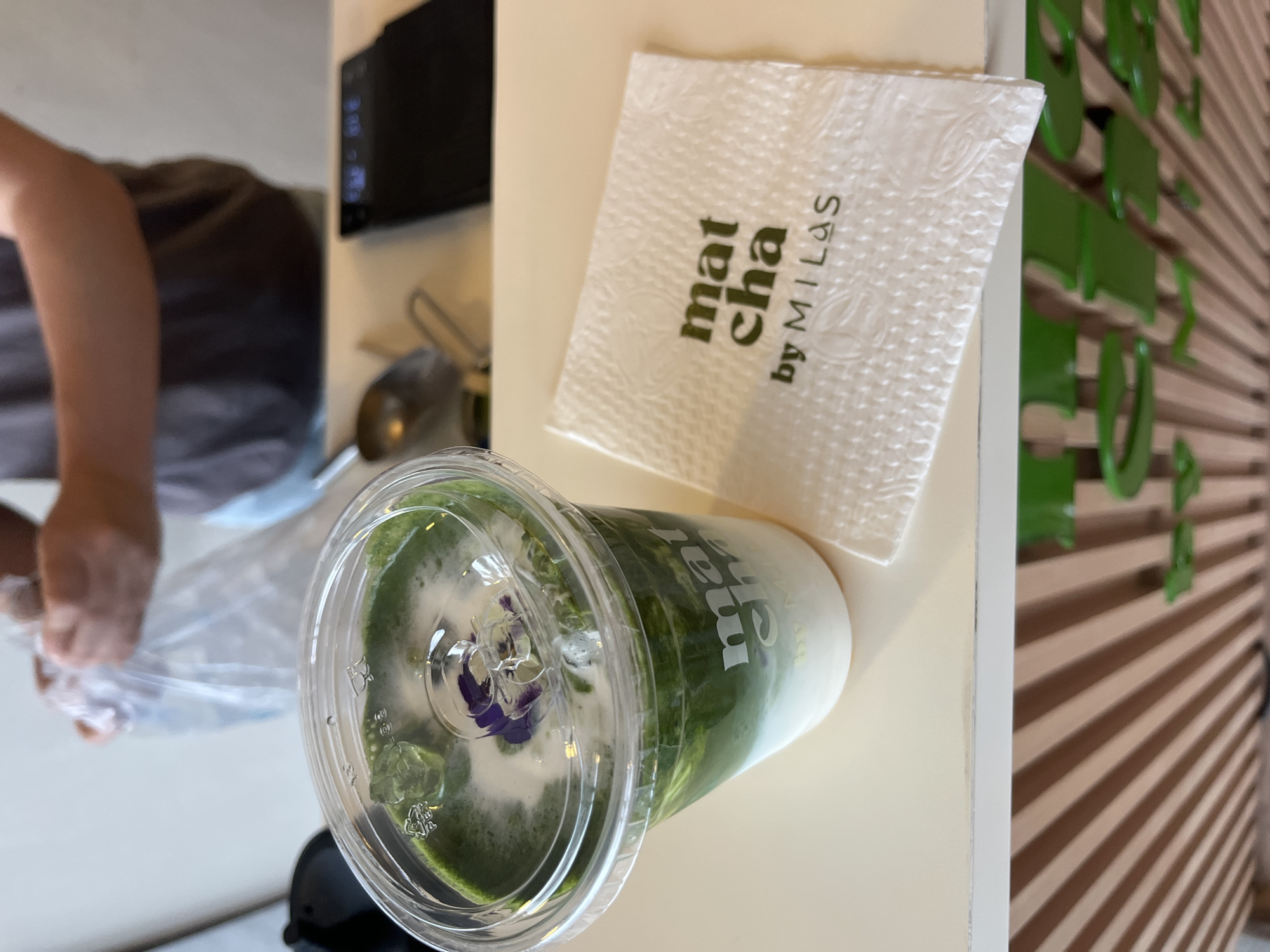 The image captures an indoor scene, likely in a cafe or a home environment in San Bernardino Tlaxcalancingo, Mexico. The central focus is a matcha drink in a clear plastic cup, placed on a light-colored countertop.
The matcha drink displays vibrant green layers at the bottom, transitioning into a creamy white layer above, suggesting a layered matcha latte. The drink is topped with a frothy white foam and a delicate purple flower garnish, possibly a butterfly pea flower, adding a decorative touch.
Adjacent to the drink is a white paper napkin, neatly folded, with visible text printed in green: "mat cha by MILAS". The napkin has a textured pattern.
In the blurred background to the left, a person is partially visible, primarily their arm and torso. They appear to be handling a transparent plastic bag. Further back, a dark appliance, possibly a microwave, is visible on a shelf. To the right, a wall features a decorative pattern of horizontal wooden slats interspersed with prominent green, possibly letter-shaped, elements, contributing to a modern or themed ambiance.