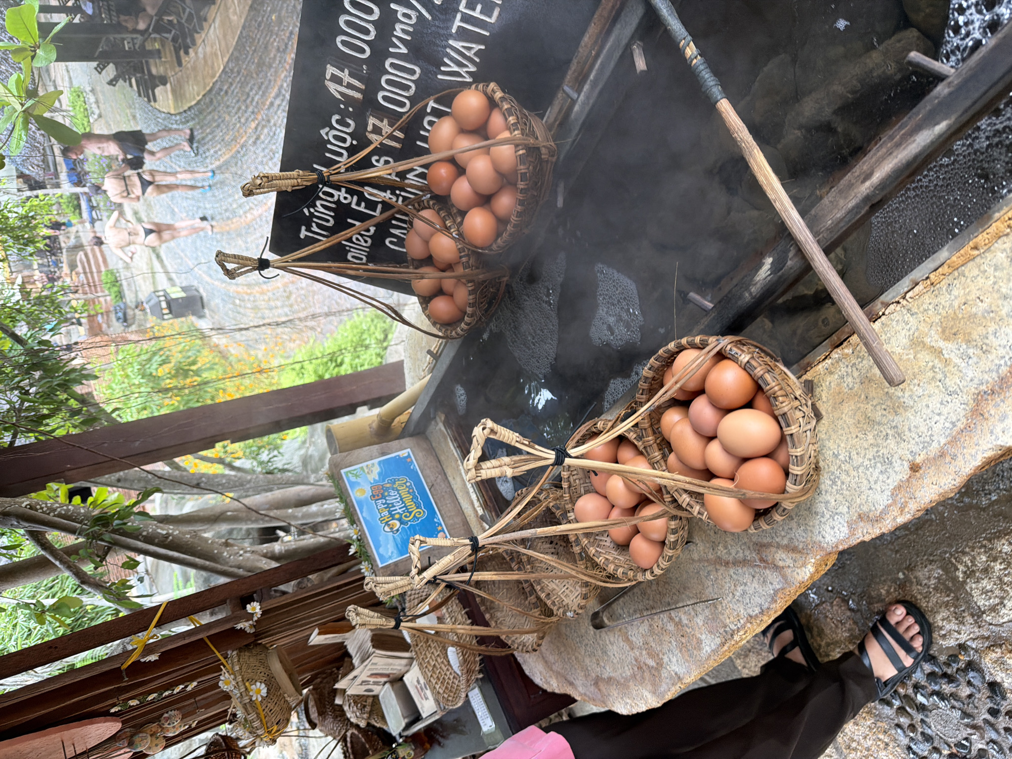 In Nha Trang, Vietnam, a steaming hot spring facility features a rustic display of boiled eggs. Three woven baskets, each suspended by a wooden handle and rope, are filled with numerous brown eggs. These baskets are positioned over a dark, bubbling body of water, indicative of a hot spring, with steam rising from the surface. A wooden tool rests across the hot spring. In the background, a chalkboard sign displays pricing in Vietnamese, listing "Trứng Luộc" (boiled eggs) at 17,000 VND, alongside "WATER". Further back, a pathway leads to a pool area where several individuals in swimwear are visible, some relaxing on loungers and others near the water, suggesting a resort or recreational setting. The overall environment appears to be outdoors, with lush greenery and trees surrounding the area.