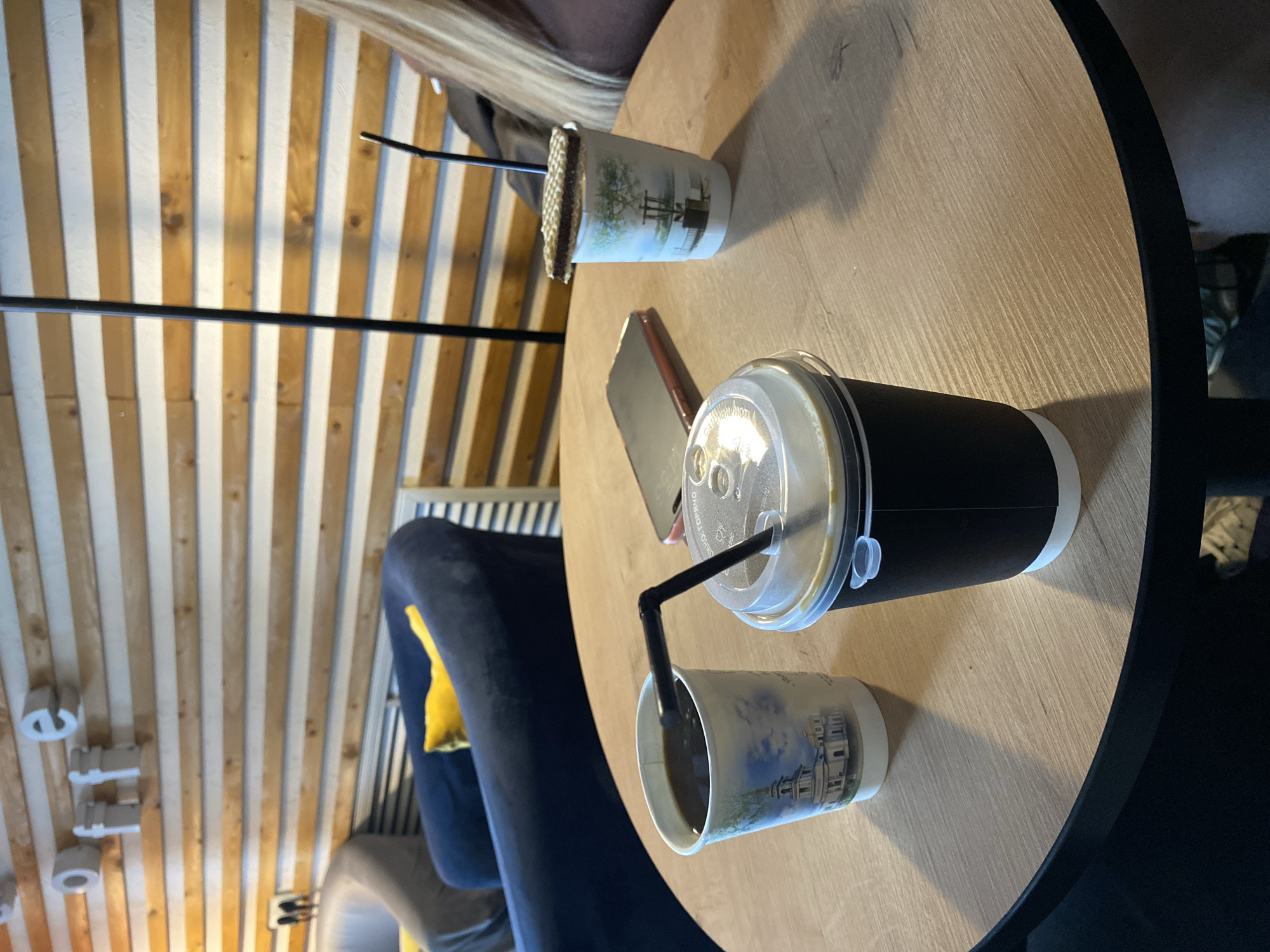 On a light wooden table, two cups of coffee sit next to a phone. One cup has a lid and a black straw, while the other has a straw and a decorative sleeve. The table is located in a cafe, with striped wooden walls and modern lighting fixtures. A blue velvet chair is visible near the table. The location is Votkinsk, Russia.