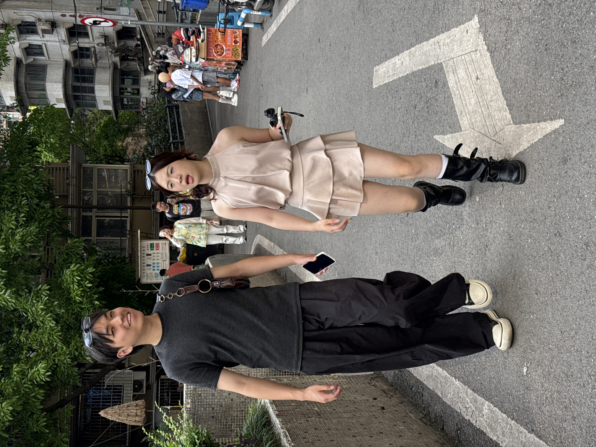 Two people, a woman and a man, stand on a city street in Yuzhong District, China. The woman wears a light-colored dress and black boots and holds a pair of sunglasses. The man wears a dark t-shirt, black pants, and white shoes. He holds a smartphone in his hand. A white arrow is painted on the street. Other people are in the background near a food vendor stand. Buildings are on the side of the street with trees and greenery. A street sign is visible.