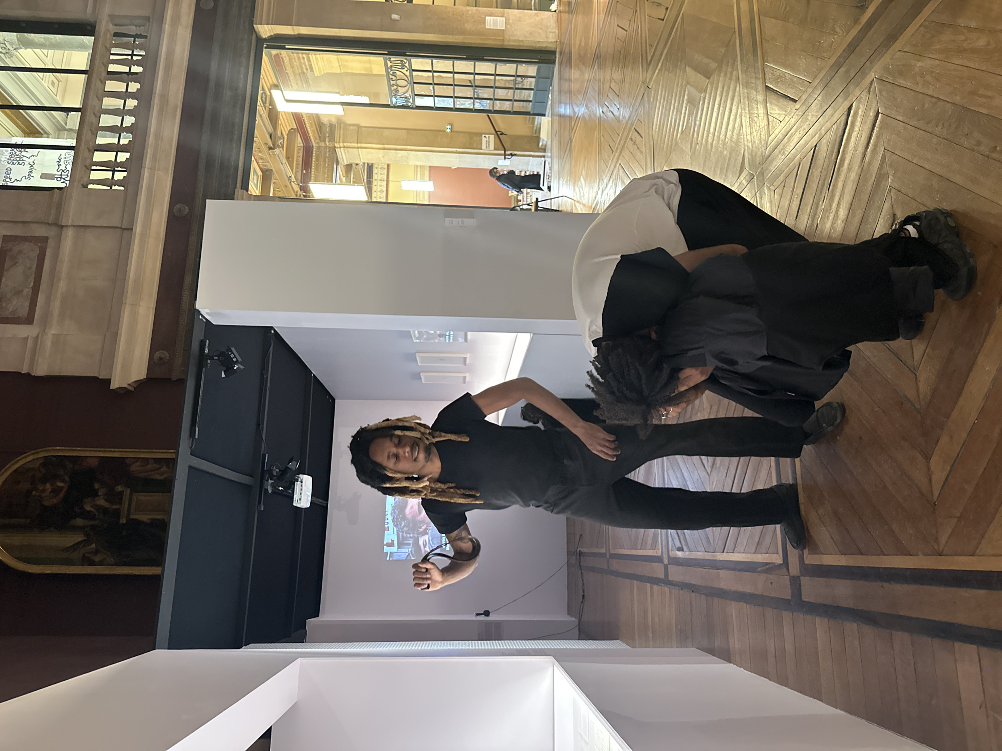 This media file captures an indoor scene within what appears to be a museum or art gallery in Paris, characterized by its ornate wooden parquet flooring and classical architectural elements. Two individuals with dreadlocks are central to the image.
One person, wearing a black t-shirt and black pants, stands towards the left, looking towards the viewer with a slight smile. They are holding a dark strap or cable in their right hand. Behind and slightly to their right, a second individual is bent over, facing downwards, dressed in a white top over black attire. Their face is not clearly visible.
The immediate surroundings include contemporary exhibition partitions in white and dark grey/black. A projector casts an image onto a white wall behind the standing person. In the background, through an open ornate doorway, a corridor leads to another person standing further away. Large windows to the left reveal more of the building's architecture, and handwritten text is visible on a surface beyond the window, including words like "Greek," "Speed," "Spank," and "RTS-Salut." A framed painting hangs on a dark wall to the far left. The lighting is predominantly artificial, supplemented by some natural light from the distant windows. The overall atmosphere is calm, suggesting an exhibition or installation viewing.