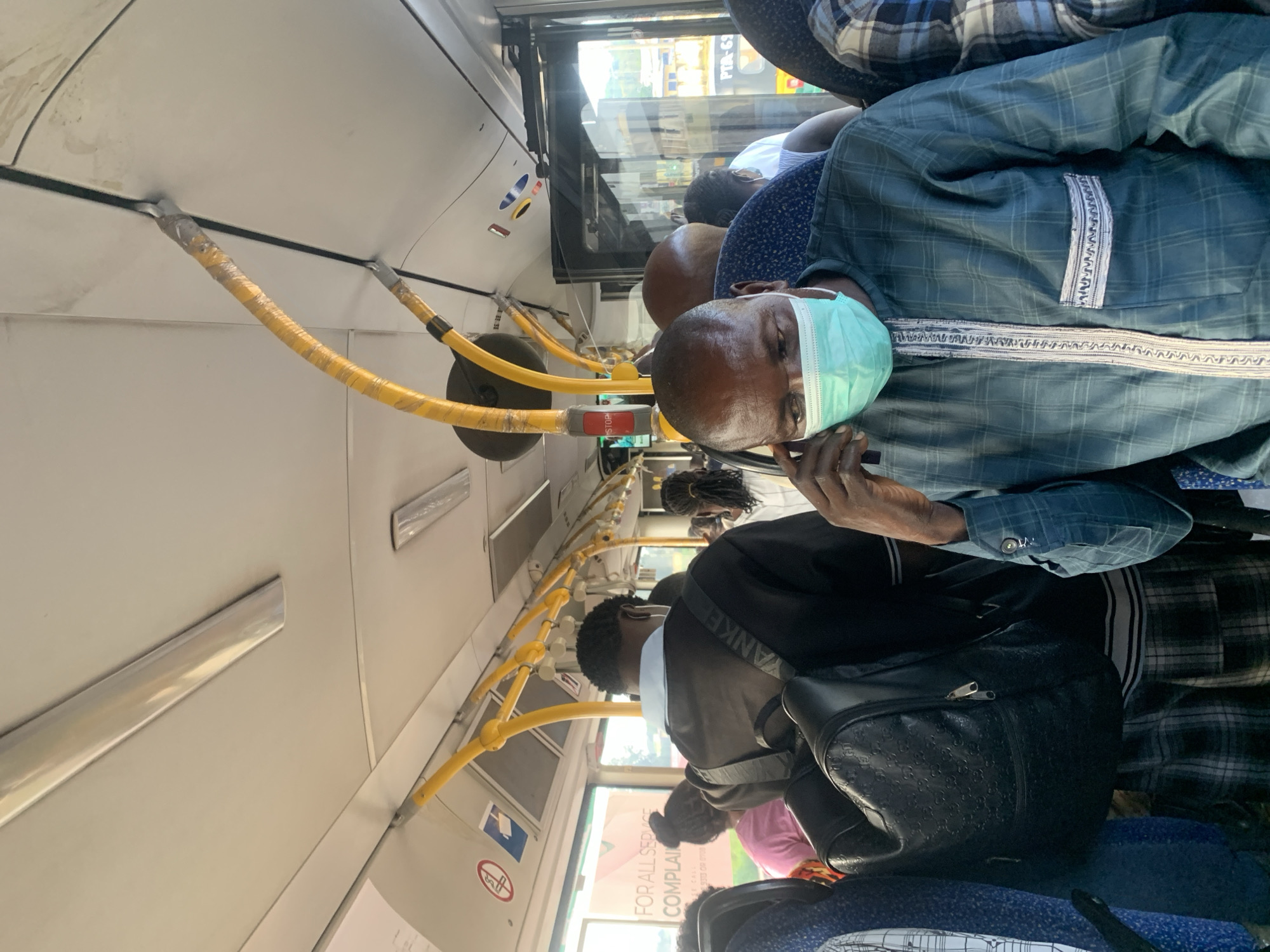 An interior view of a crowded bus, featuring a man wearing a light blue face mask and a dark teal plaid long-sleeved shirt with ornate trim and a chest pocket. He is holding his right hand to his ear, as if on a phone call. The man is seated, and the focus is on his face and upper body. In the background, other passengers are visible, some wearing masks, and the bus interior includes yellow grab poles, ceiling lights, and windows. The scene appears to be within a city.