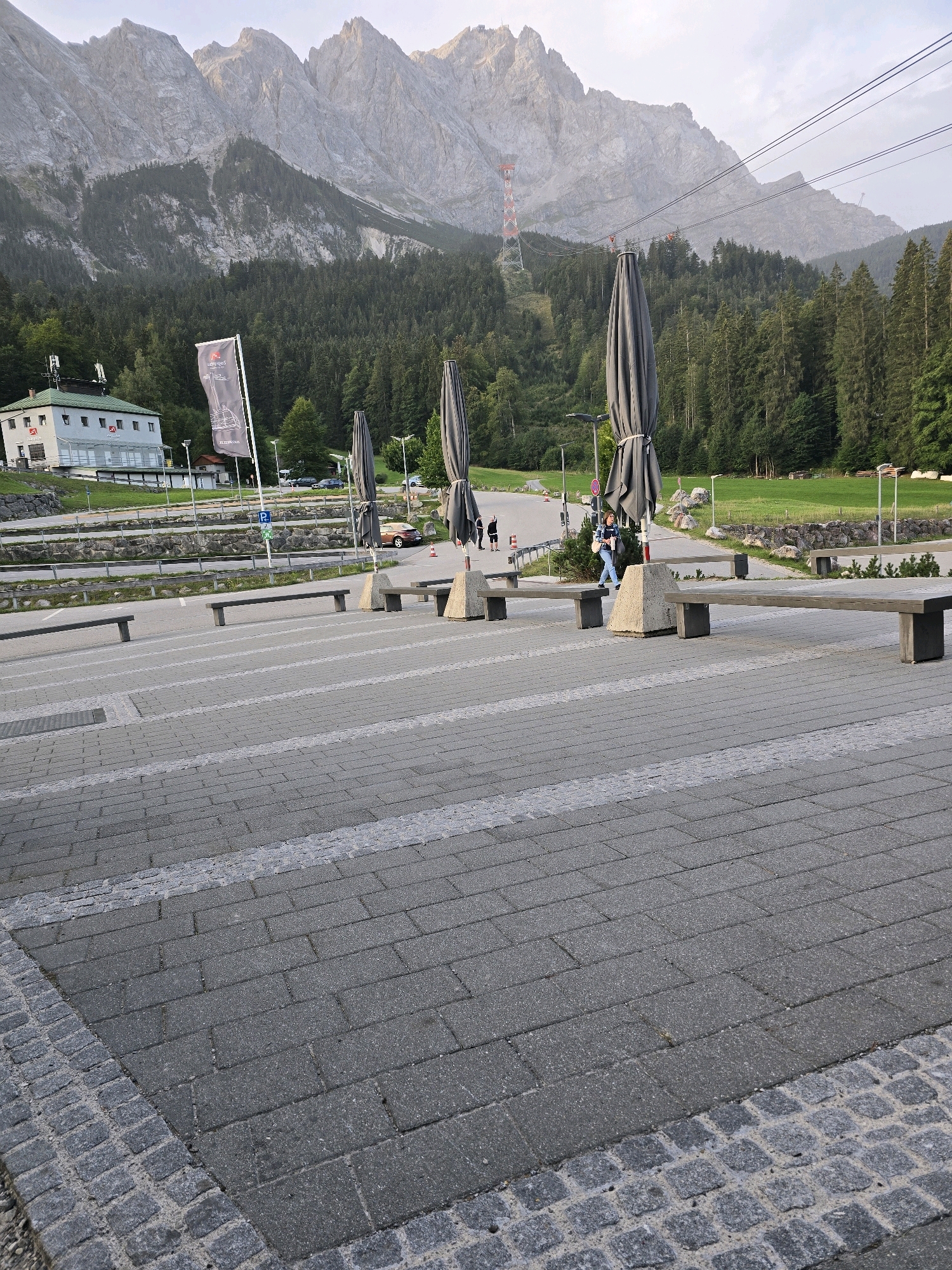 In Grainau, Germany, this image captures a vast, tiered plaza paved with dark gray interlocking cobblestones, accented by lighter gray, linear stone inlays. Several long, dark gray benches with concrete bases are strategically placed across the plaza. To the left, a modern, light-colored building with a green roof and a red accent on its facade stands adjacent to a grey flagpole bearing a black logo. In the mid-ground, a paved road runs horizontally, bordered by a low stone wall. Several cars are parked along this road, and a blue parking sign is visible. Three large, closed dark gray patio umbrellas are erected on the plaza, flanking the road. A woman in blue jeans and a white and blue patterned shirt is walking on the plaza, looking down at her phone. In the background, a dense forest of dark green pine trees covers the lower slopes of a majestic, jagged mountain range, whose peaks are illuminated by the soft light of what appears to be late afternoon or early morning. A prominent red and white radio tower is visible on one of the mountain slopes, alongside a cable car system with its cables stretching across the scene. The sky above is a pale, hazy blue.