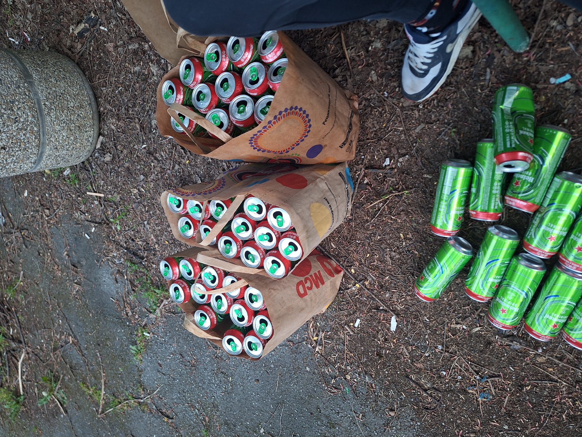 The scene depicts a collection of empty aluminum cans, mostly Heineken and another brand, scattered on a ground covered in leaves and debris, likely in an outdoor environment. Three brown paper bags, printed with "McD" and other logos, are filled with the red-and-green cans. A person wearing a blue and white shoe is partially visible in the upper right corner of the frame. A concrete cylindrical object is located in the upper left of the frame, along with what appears to be a green cylindrical object. The image was taken in Sofia, Bulgaria, due to the Cyrillic writing on the brown paper bag.