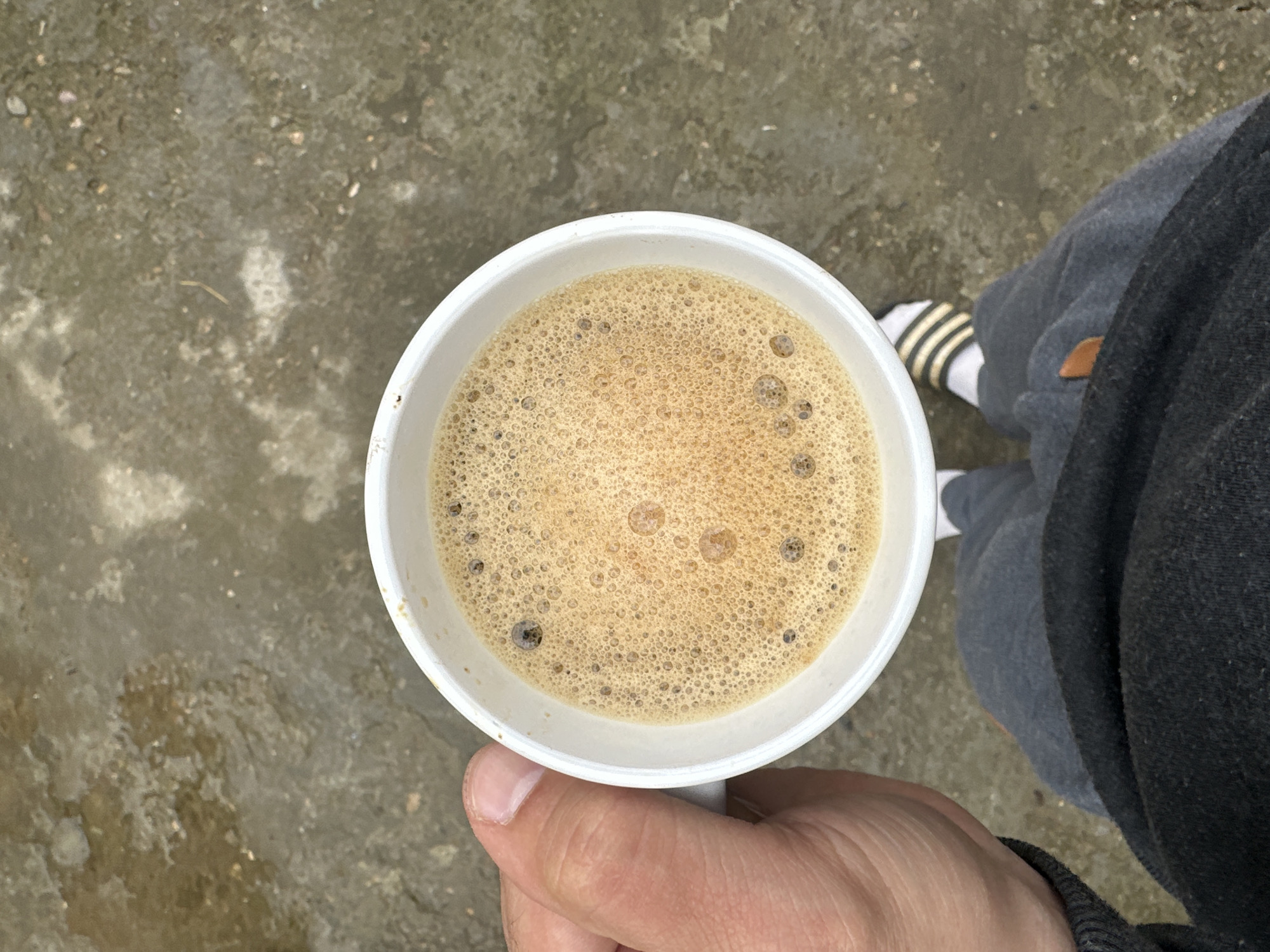 A top-down view shows a hand holding a white foam cup filled with a light brown foamy liquid, likely coffee or a similar beverage. In the background, the ground appears to be wet concrete or stone. To the right, a person's legs are visible, wearing grey sweatpants, white socks with black stripes, and striped sandals. The photo was not taken in Slavyani, Bulgaria.