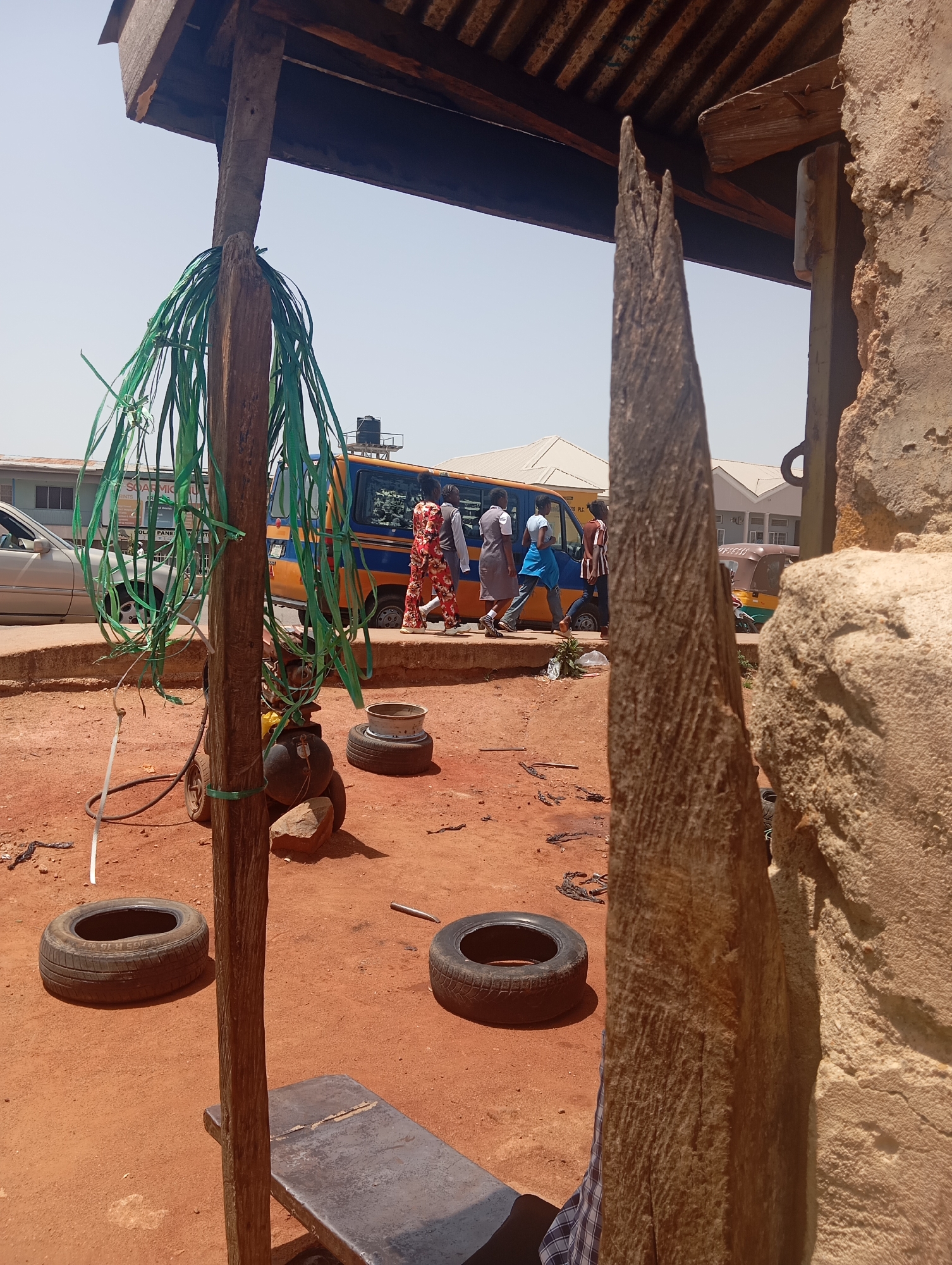 This media file captures an outdoor scene in Jos, Nigeria, with a focus on a rustic setup that appears to be a tire repair or sales area. The foreground is dominated by a rough wooden pole with green plastic strapping wound around it, hinting at repurposed materials. Several tires are visible, some stacked and others lying flat on the reddish-brown earth, suggesting their purpose as either spare parts or for sale. A metal bench or scale is positioned near the base of the pole.
In the background, a yellow and blue mini-bus is parked, indicating a transportation hub or a roadside service area. Several people are walking or standing near the bus, some appearing to be customers or workers. One woman is wearing a vibrant, patterned red and white outfit, while others are dressed in more casual attire. A small, multi-colored auto-rickshaw is also parked further in the background, a common mode of transport in the region. The architecture of the buildings in the distance is simple, with light-colored facades and visible roofs.
The overall impression is one of a bustling, everyday scene in an urban or semi-urban area. The bright sunlight suggests it is daytime, likely a warm, clear day. The ground is dusty red earth, typical of many parts of Nigeria. There are no overt emotional cues, but the general atmosphere feels active and functional. Some text is visible on the side of the mini-bus, but it is too blurry to be legible. The scene conveys a sense of local commerce and daily life.