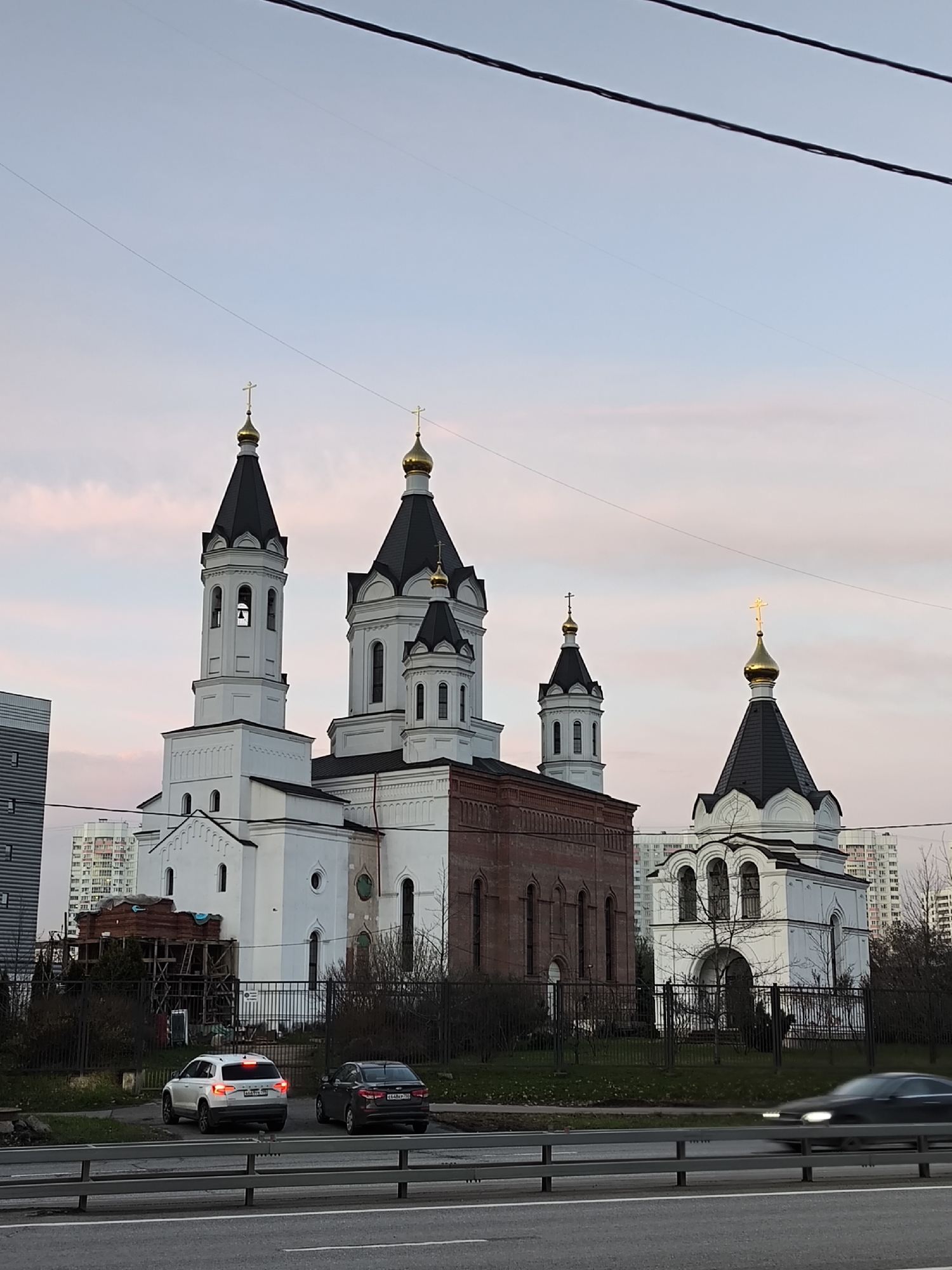 This evening urban scene captures a large Orthodox church complex under a sky painted with soft pink, orange, and light blue hues of dusk. The central structure is a multi-domed church, predominantly white with sections of red brick, featuring black conical roofs and shimmering golden onion domes, each topped with an Orthodox cross. A tall bell tower, also white with a black roof and golden cross, stands prominently to the left. To the right, a smaller, entirely white chapel-like building with a similar architectural style and a golden-domed cross completes the complex. The church grounds are bordered by a black metal fence, with bare trees and some construction scaffolding visible near the church's base.
In the background, numerous multi-story residential apartment buildings and a modern, dark-colored building on the far left suggest a dense urban setting.
The foreground shows a multi-lane asphalt road separated by a grey guardrail. Two cars are parked or moving slowly on a side road near the church fence: a white SUV (license plate M449MM 190) and a dark sedan (license plate E480AE 790). Another dark sedan is captured in motion on the main road, appearing blurred. Overhead, dark power lines stretch across the sky. The scene depicts a tranquil moment in Moscow, Russia, as artificial lights begin to complement the fading natural light.
