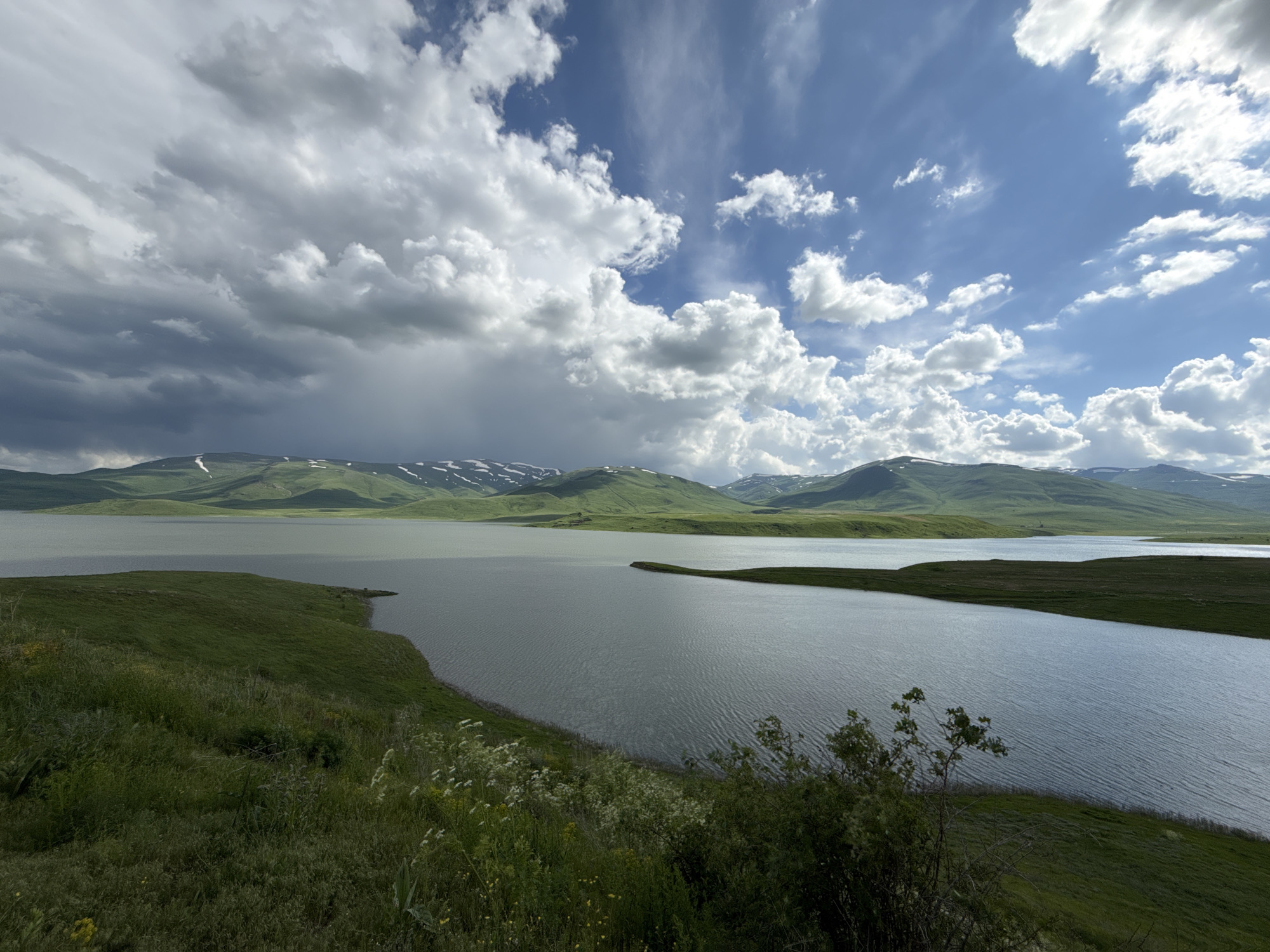 The image shows a body of water with rolling green hills in the background, some of which are capped with snow. The sky is a mix of blue with white and gray clouds, suggesting variable weather. The foreground consists of a grassy slope with scattered wildflowers and some low-lying bushes. The scene is a natural landscape with no people or man-made structures visible. This landscape is characteristic of the mountainous regions of Armenia, including the area around Tsghuk.