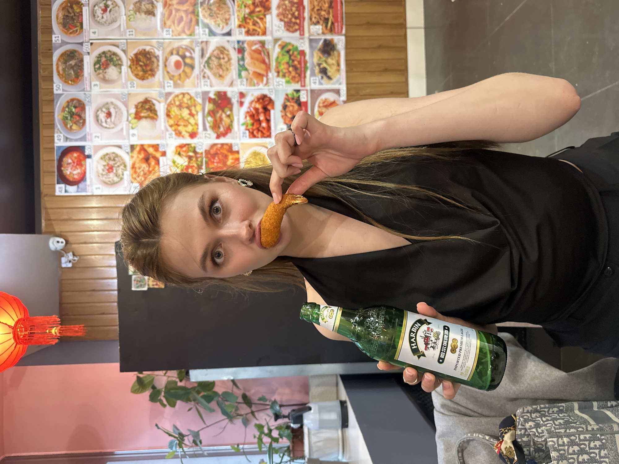 A young woman is in a restaurant, holding a bottle of Harbin beer and eating a fried shrimp. Behind her is a wall with a menu of various dishes. The restaurant might be located in the city of Krasnoyarsk, Russia. She is wearing a black top and has a handbag on the seat beside her. There's a plant and a red lantern in the background.