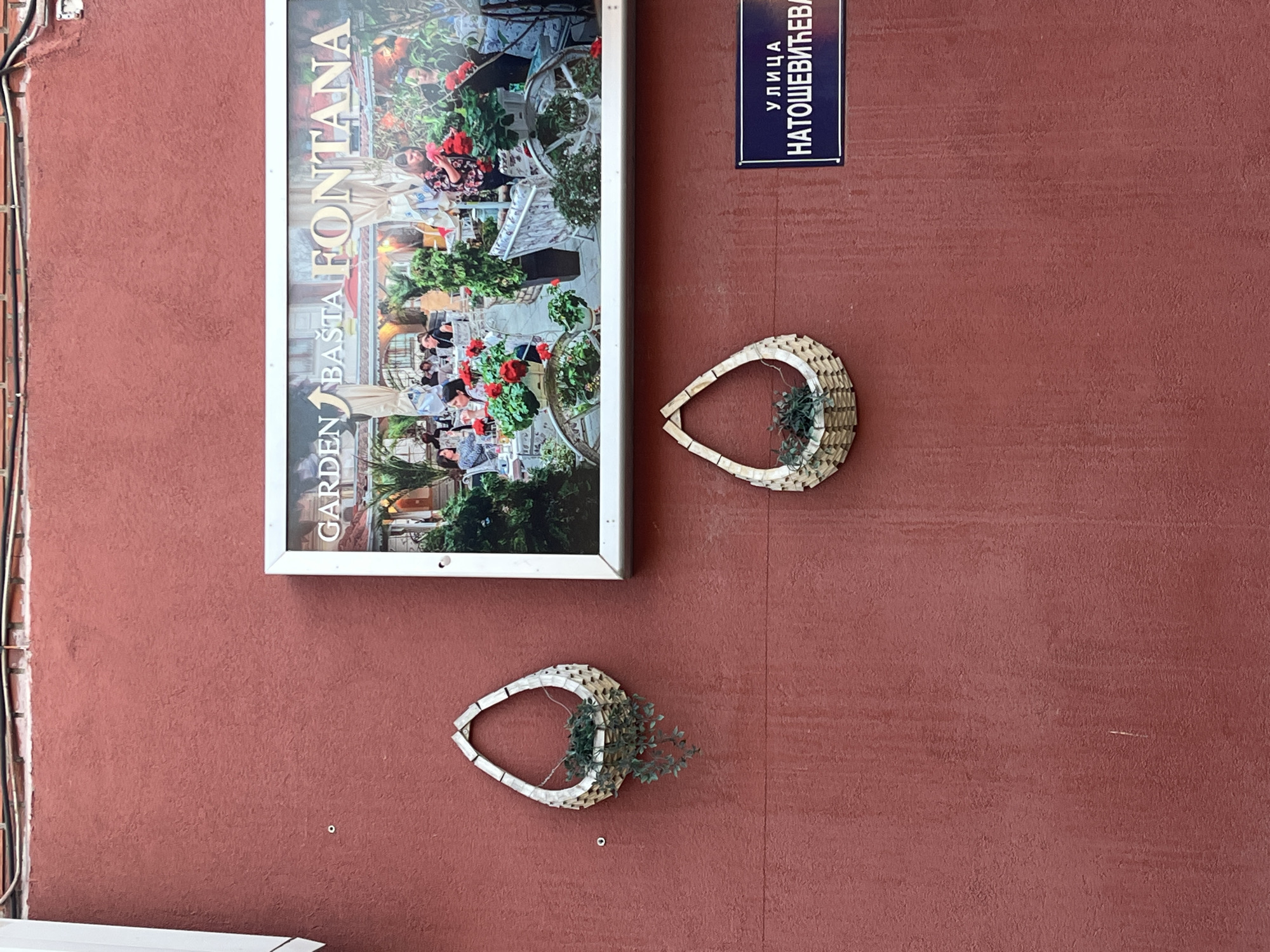 The image shows a wall in Novi Sad, Serbia. On the wall, there's an advertisement for "GARDEN BASTA FONTANA" featuring a scene of people dining outdoors amidst lush greenery. Above the advertisement, there is a blue street sign reading "улица НАТОШЕВИЋЕВА" (Natoševićeva Street). Below the sign and advertisement, there are two decorative woven baskets, each containing some greenery, affixed to the wall. The wall is painted a terracotta-red color. A portion of a white door frame is visible on the bottom left of the image.