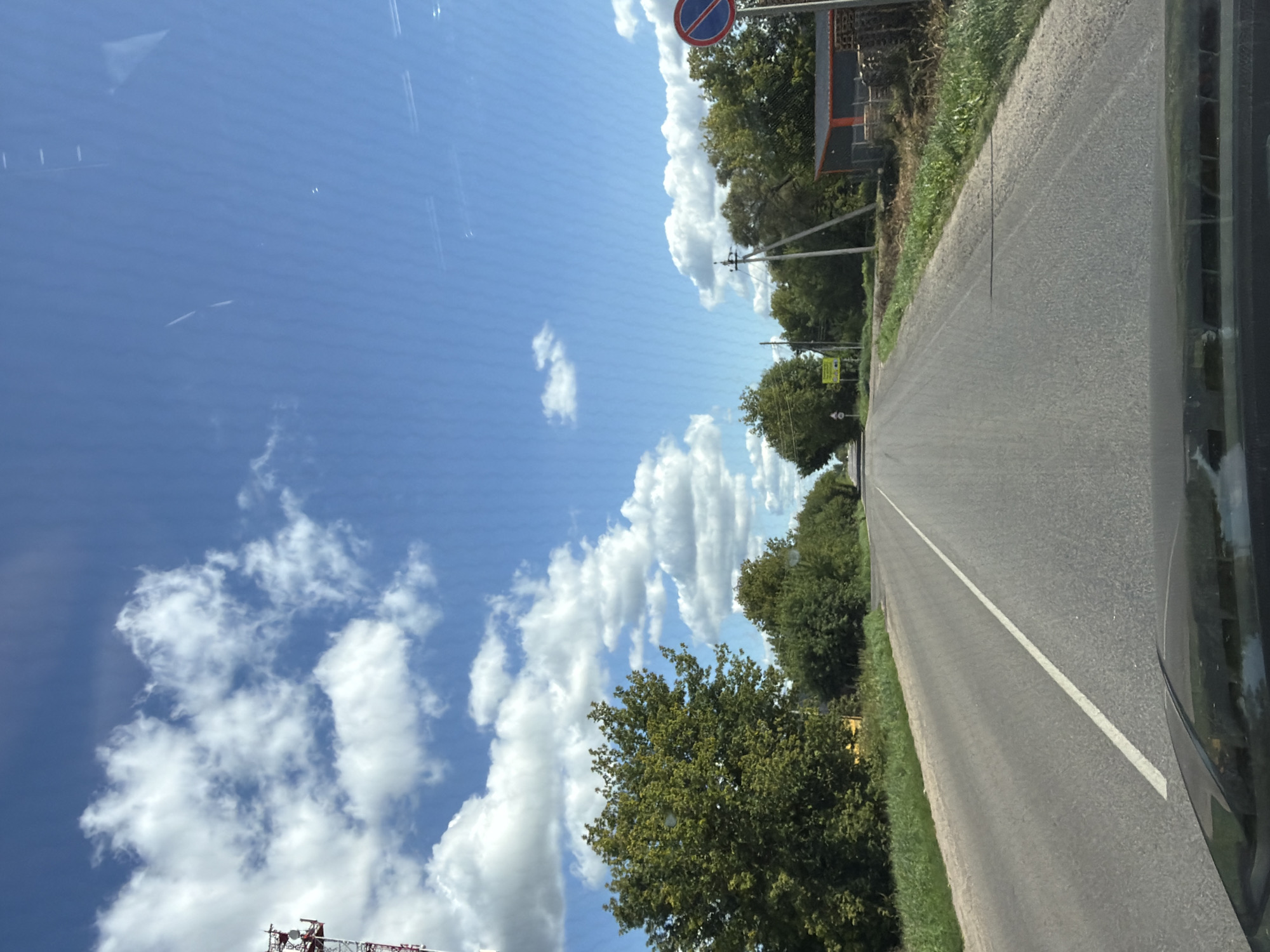 The image captures a sun-drenched asphalt road leading into the distance, bordered by lush green trees and vegetation. The sky is a vibrant blue, dotted with fluffy white clouds. On the left side of the road, a tall red and white communication tower stands prominently. Further down the road, a small yellow sign is visible. On the right side, a small orange and gray building with stacked wooden pallets in front of it is present. A circular traffic sign with a red border and a blue diagonal bar inside is also visible on the right. The perspective suggests the photo was taken from inside a vehicle, looking forward. This scene is located in Akseno-Butyrki, Russia.