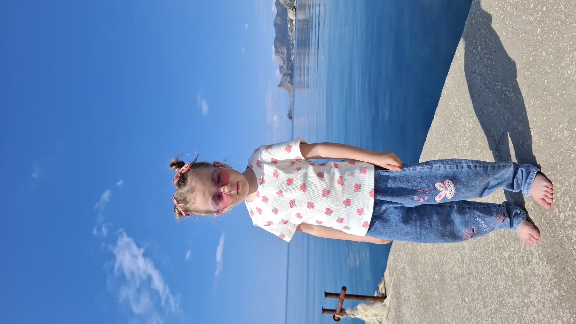 A full shot, rotated counter-clockwise, presents a young girl standing on a concrete pier or dock. The girl has blonde hair styled in two small buns, each adorned with pink bows. She wears pink sunglasses, a white t-shirt with a floral pattern, and blue jeans embellished with bunny designs.
The background consists of a vivid blue sky with scattered clouds and the ocean. Distant mountains can be seen across the water. The pier is marked with a rusted, metal structure near the edge. The location is Unknown, Unknown.