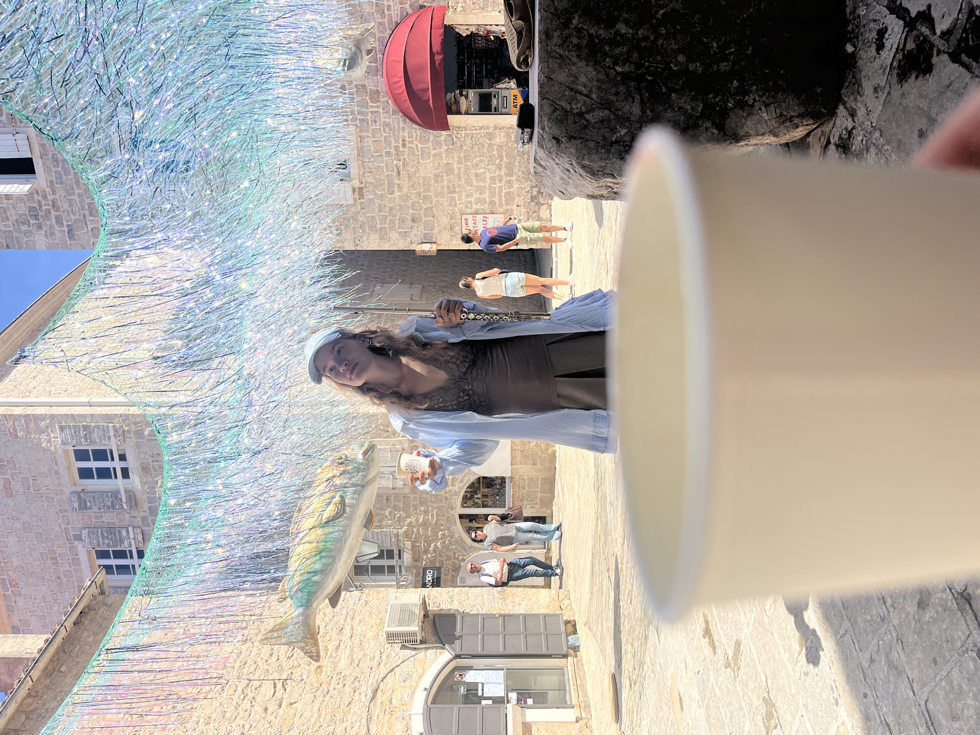 The image shows a woman standing in a stone-paved street in what appears to be the old town of Budva, Montenegro, holding a cup in one hand and a long flute in the other. The foreground is dominated by an out-of-focus white cup, obscuring part of the scene.
Above the street are iridescent decorations resembling a fishing net with a large ornamental fish hanging from it. Buildings on either side of the street are constructed of stone. Various shops and doorways line the street, and several pedestrians are visible in the background. The architecture suggests a historic Mediterranean setting. The street is narrow, and the angle of the shot is upwards, capturing the decorations above.