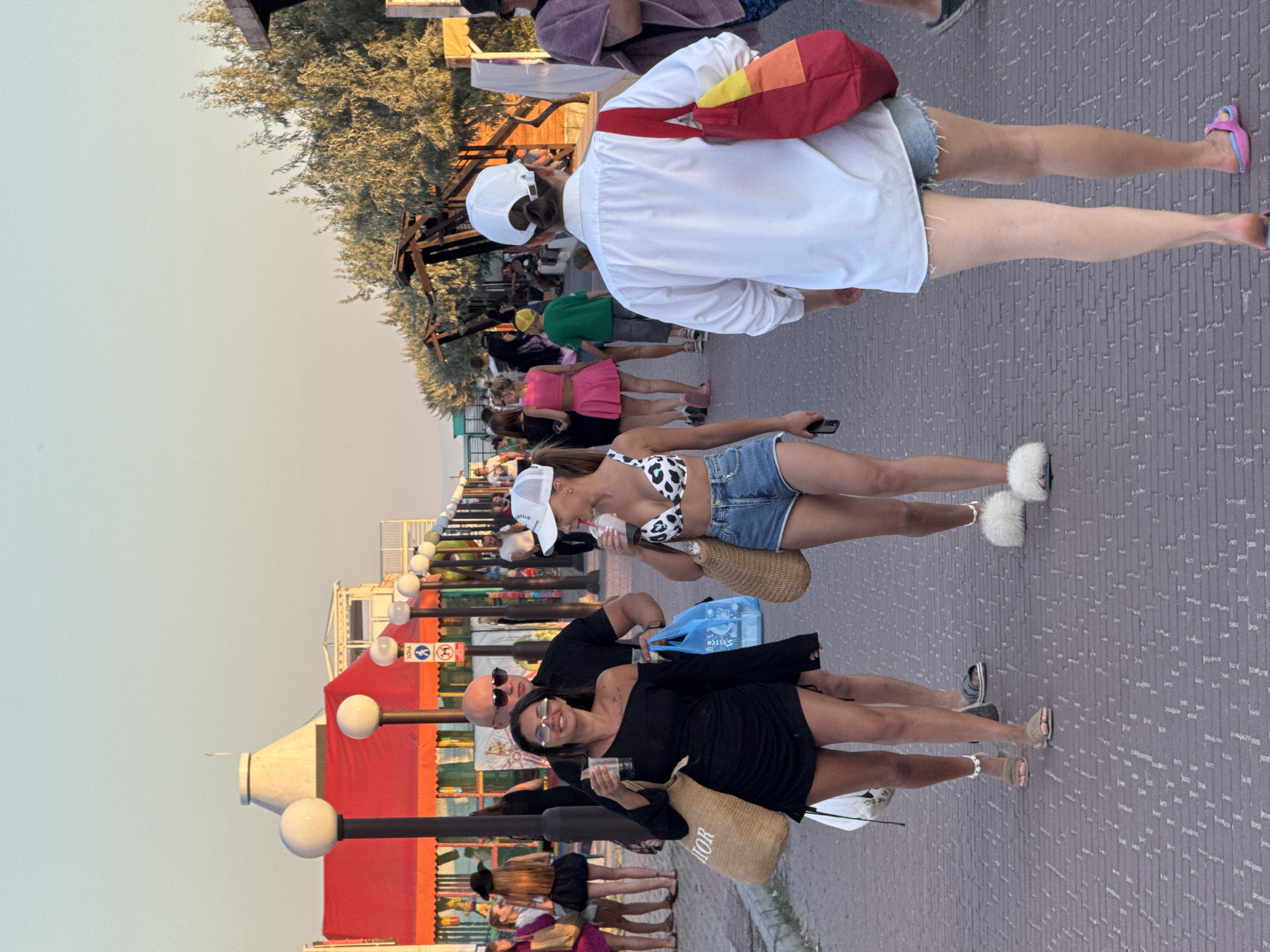 This image depicts a street scene in Novofedorovka, Ukraine, under a clear, pale sky, possibly during late afternoon or early evening, with the sun casting a warm glow. The foreground shows a paved walkway made of brick-like material.
Several individuals are walking along the path. On the left, a woman in a black off-the-shoulder dress and sunglasses is smiling and holding a clear plastic cup. She carries a large, woven tote bag over her left shoulder and a blue plastic bag in her right hand. Beside her walks a man in a black t-shirt and sunglasses.
Further ahead and slightly to the right, another woman in a white baseball cap, a leopard-print bikini top, and denim shorts is walking and holding a clear plastic cup. She has a straw tote bag over her shoulder. Behind her, and to the right of the frame, a person wearing a white button-down shirt, denim shorts, and a white baseball cap walks away from the camera, with a red, yellow, and orange tote bag slung over their left shoulder.
In the background, a red awning covers a structure with several lamps on poles lining the walkway. Further back, there are trees and what appear to be market stalls or kiosks. Other people are visible in the mid-ground and background, some appearing to be seated. The overall atmosphere suggests a lively pedestrian area, likely near a beach or resort.