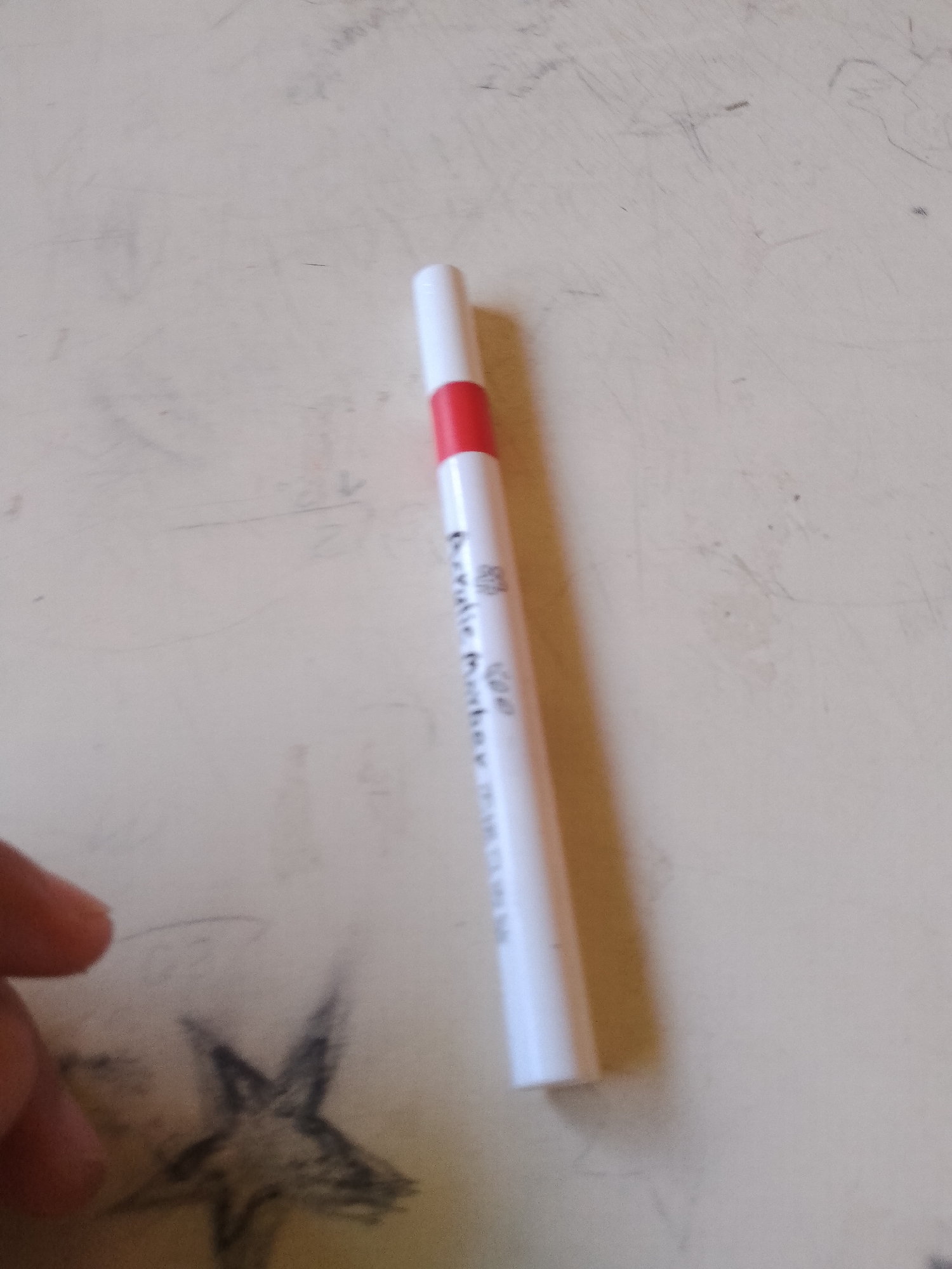 The image shows a white and red marker pen lying on a textured, light-colored surface. The pen has black text printed vertically on its barrel, which is partially legible and appears to read "Dry-Erase Marker". To the left of the pen, a blurred image of a hand is visible, as is a faint sketch of a star. The background is a plain, lightly textured surface, possibly a desk or table. There are some faint lines and smudges on the surface, indicating it may have been used for writing or drawing. The lighting is relatively even, suggesting an indoor environment. The image does not contain any specific geographical or contextual information that would allow for its location to be identified within the city of Santa Ana, Venezuela.