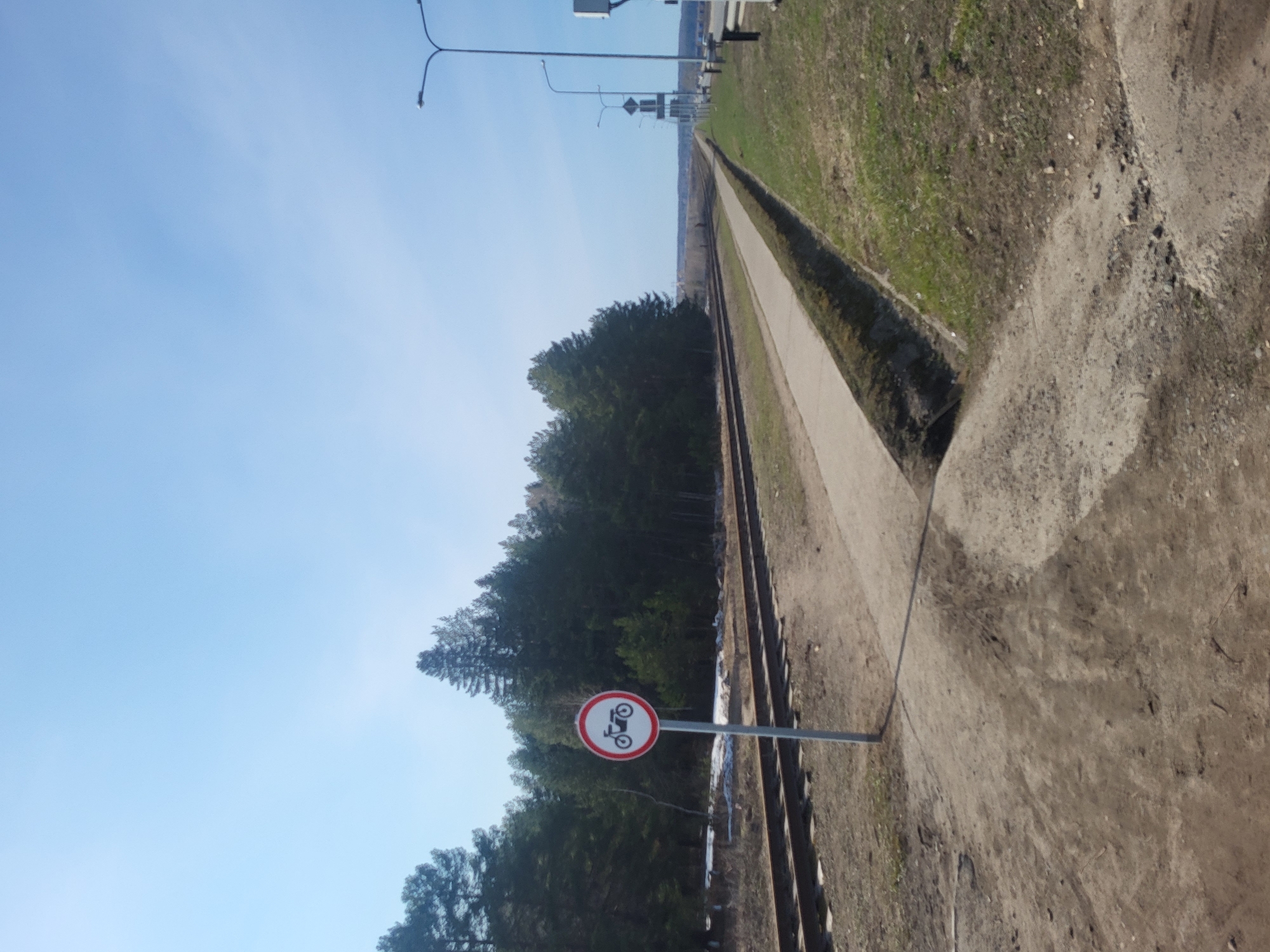 The image depicts a railway line in what appears to be a rural or semi-rural area. The railway tracks run parallel to a paved path or road. A "No Motorcycles" sign is visible along the path, indicating that motorcycles are prohibited in this area. The environment includes trees, grass, and dirt, suggesting a natural or undeveloped setting. In the background, there are some street lights and what appears to be a bridge or overpass. While the image does not contain specific details to precisely pinpoint the location, the "No Motorcycles" road sign is standard in Russia, making the city of Chaikovsky a reasonable guess.