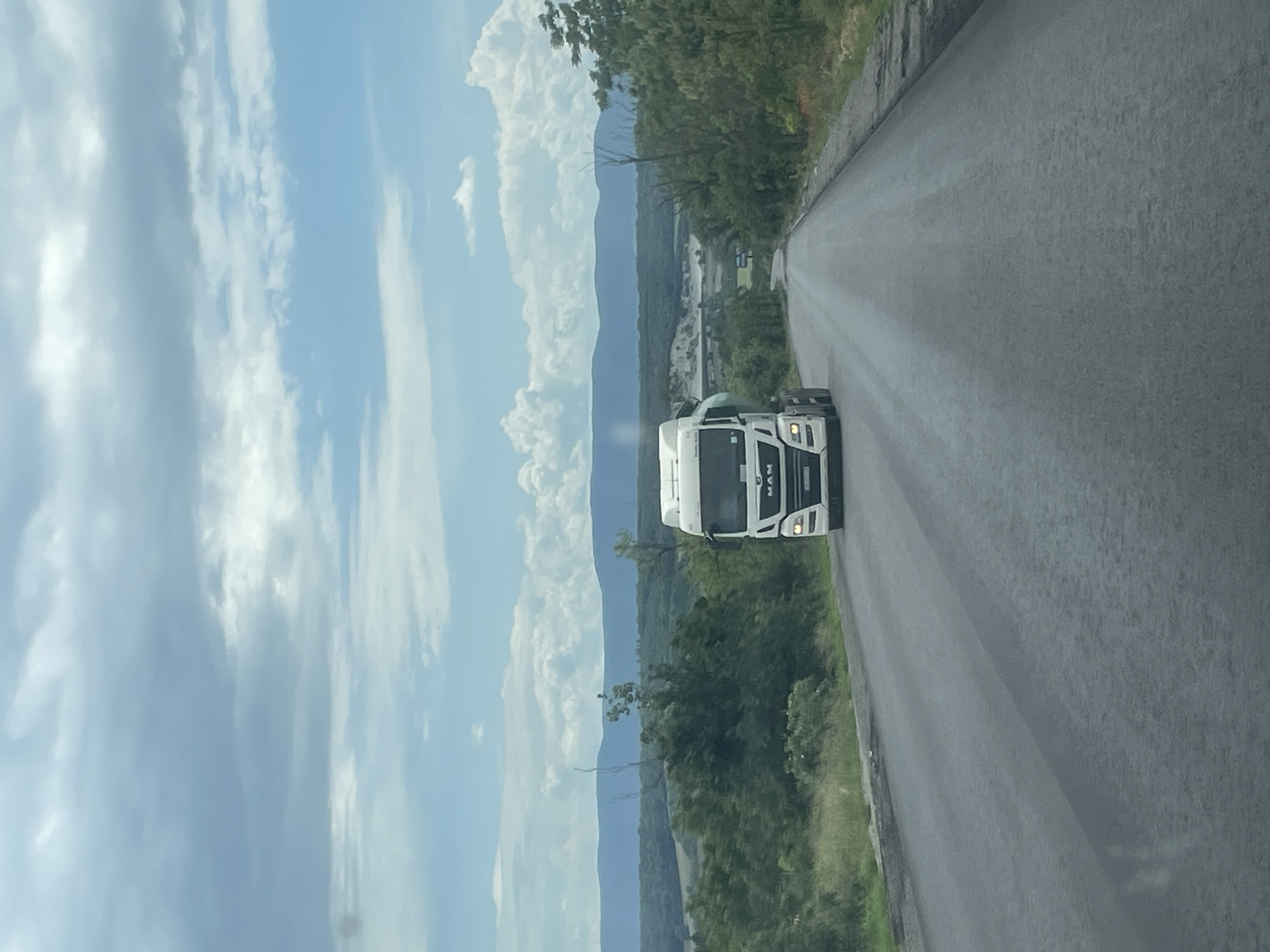 A medium shot depicts a white MAN truck traveling on a road in the vicinity of Alvanovo, Bulgaria. The sky is blue with cumulus clouds. Mountains are visible in the background, along with verdant foliage and distant structures, situated either in or near Alvanovo. The road is paved and asphalt.