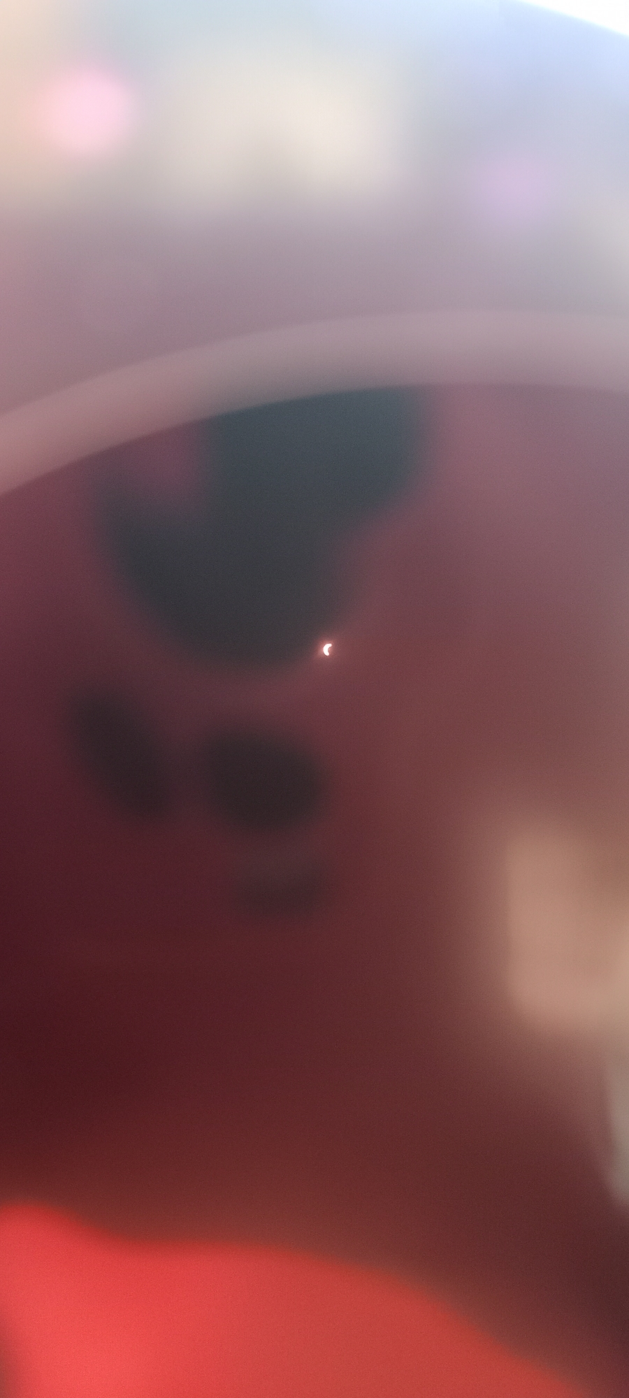 The image depicts a partially eclipsed sun, appearing as a crescent shape, against a diffuse reddish-pink background. There are some darker, amorphous shapes visible in the background, possibly clouds or obstructions. The overall image is out of focus, with a bright light source in the upper portion. There is no indication of the city of Adai, Spain, or any other specific location within the image itself.