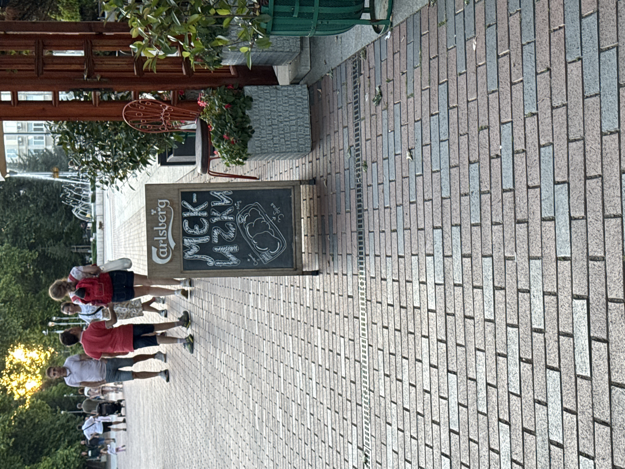 An outdoor scene in a city features a brick-paved walkway where several people are walking. The sun is setting, casting a warm glow through the trees on the left side of the frame. In the foreground, a chalkboard sign displays a menu in Cyrillic script, advertising "Мек-ички" and a drawing of a sandwich. A red chair with an ornate back and a stool are placed near the sign, along with potted plants. In the background, a fountain sprays water, and a building with multiple windows is visible.