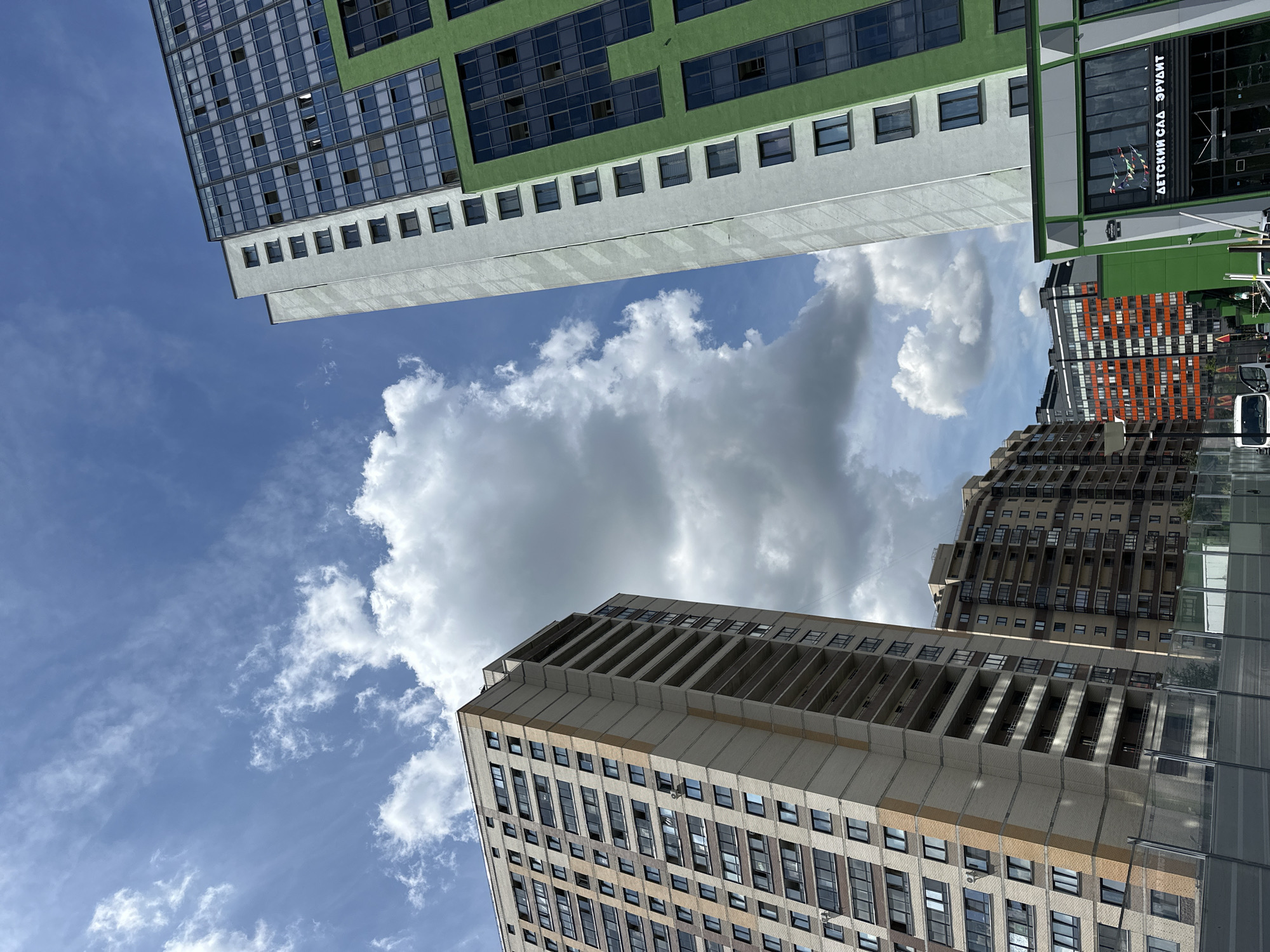 The image captures a daytime view of modern high-rise residential buildings in Kudrovo, Russia. The perspective is from a low angle, looking upwards towards the sky. In the foreground, a light-colored apartment building with numerous windows and balconies dominates the left side. To the right, a taller, more contemporary building features a striking green facade combined with white and dark-paned windows. Further in the background, other residential blocks with varying architectural styles and colors are visible, including one with orange accents. The sky is a bright blue with scattered white clouds, suggesting fair weather. The lower portion of the image shows fencing and the roof of a vehicle, implying a street-level viewpoint. One building in the foreground has a sign that reads "ДЕТСКИЙ САД ЭРУДИТ" (Children's Garden Erudite).