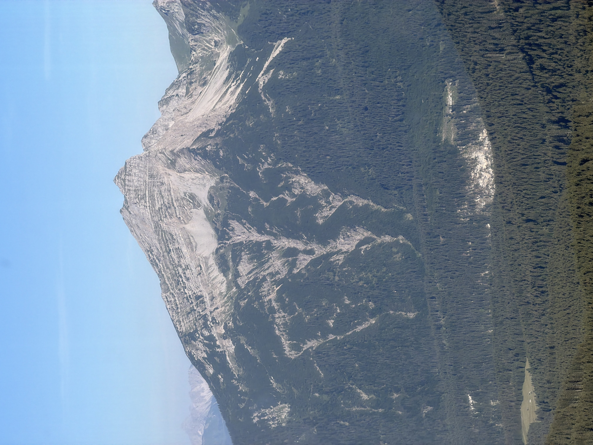This is a wide-angle, aerial photograph of a mountainous landscape under a clear blue sky, likely taken from an elevated vantage point. The foreground is dominated by a dense, dark green coniferous forest. In the mid-ground, the slopes of the mountains are covered in the same forest, with lighter-colored rock formations and scree visible in patches, particularly along the steeper inclines. These rock faces are streaked with lighter gray and white, suggesting erosion and bare rock.
The background features a prominent, jagged mountain peak, its summit appearing stony and barren, with stratified layers of rock clearly visible. The slopes leading up to this peak are a mix of green vegetation and lighter rock. Further in the distance, additional mountain ranges are visible, appearing softer and more hazy due to atmospheric perspective, with peaks also exhibiting lighter coloration.
In the lower left portion of the image, nestled within the forest, is a small, grassy clearing. Within this clearing, there are at least three small, light-colored structures, resembling buildings or cabins, possibly part of a small settlement or a secluded farm. The presence of these structures, combined with the mountainous terrain and extensive forests, is consistent with the landscape found in and around Grainau, Germany, a town known for its proximity to the Bavarian Alps. There are no people or animals visible in the scene. The overall impression is one of rugged natural beauty and a remote, undeveloped environment.